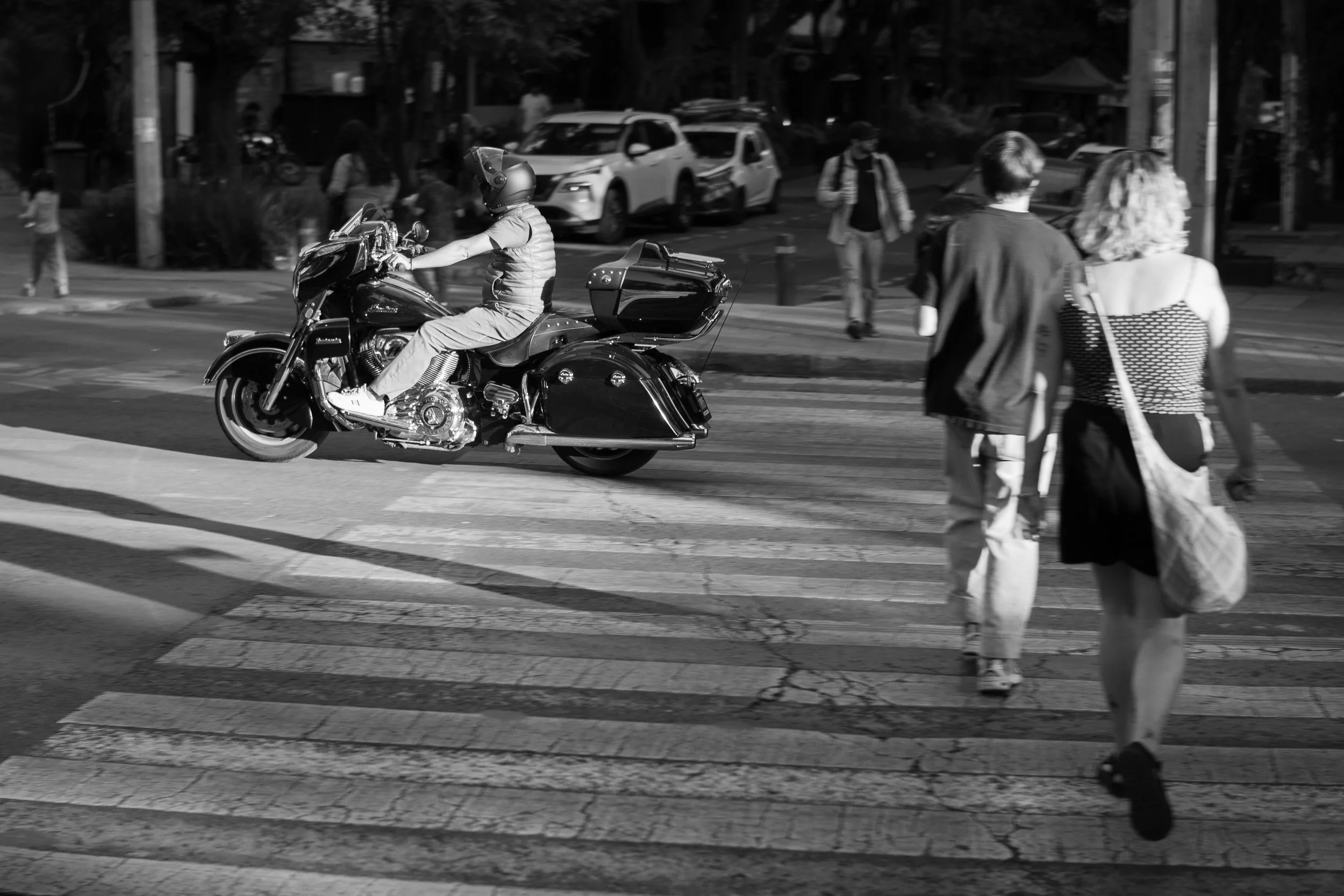 Una escena urbana en blanco y negro con una motocicleta y una pareja cruzando la calle. (c) 2025 Héctor Obregón