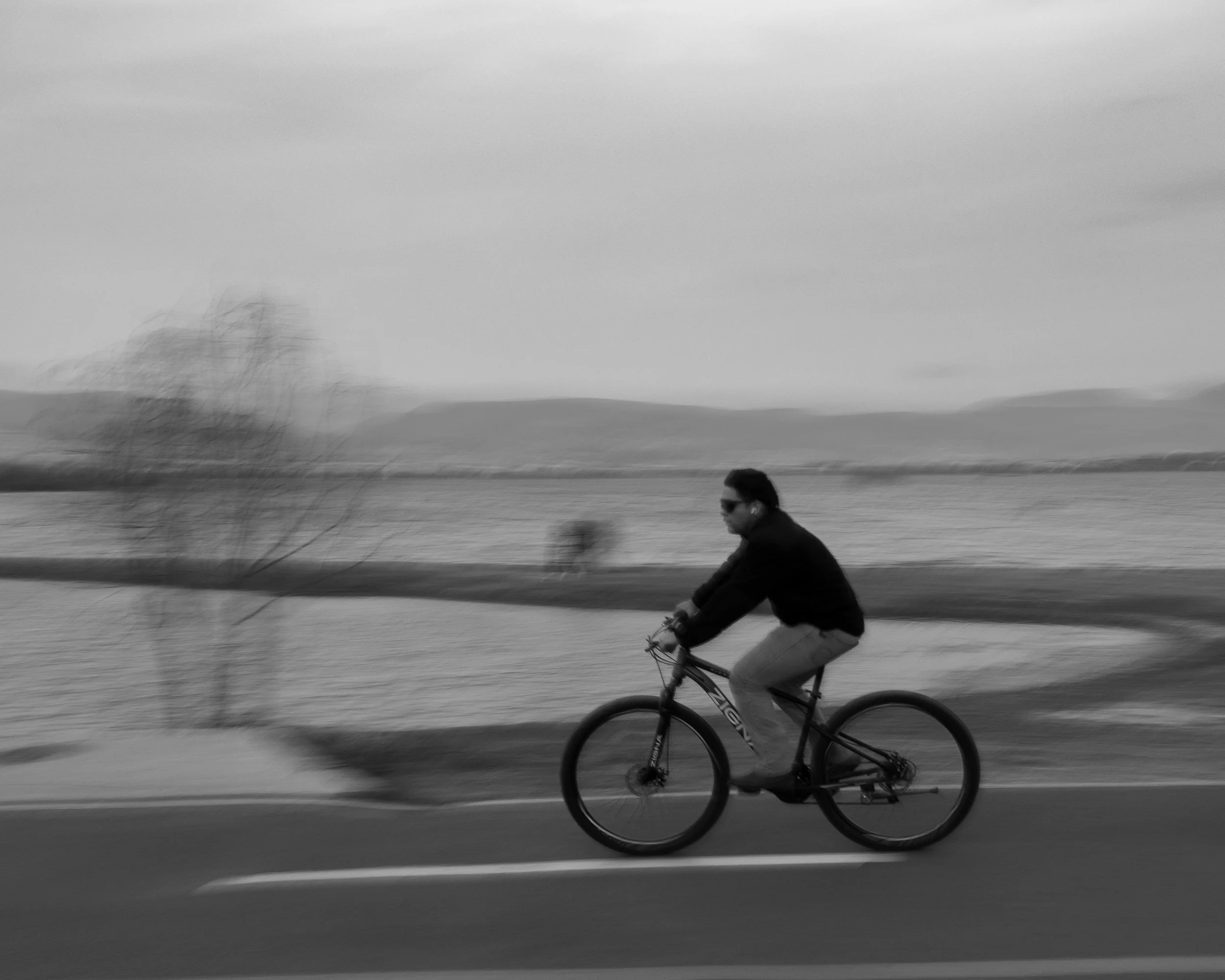 Hombre montando bicicleta en un día nublado junto a un lago. (c) 2026 Héctor Obregón