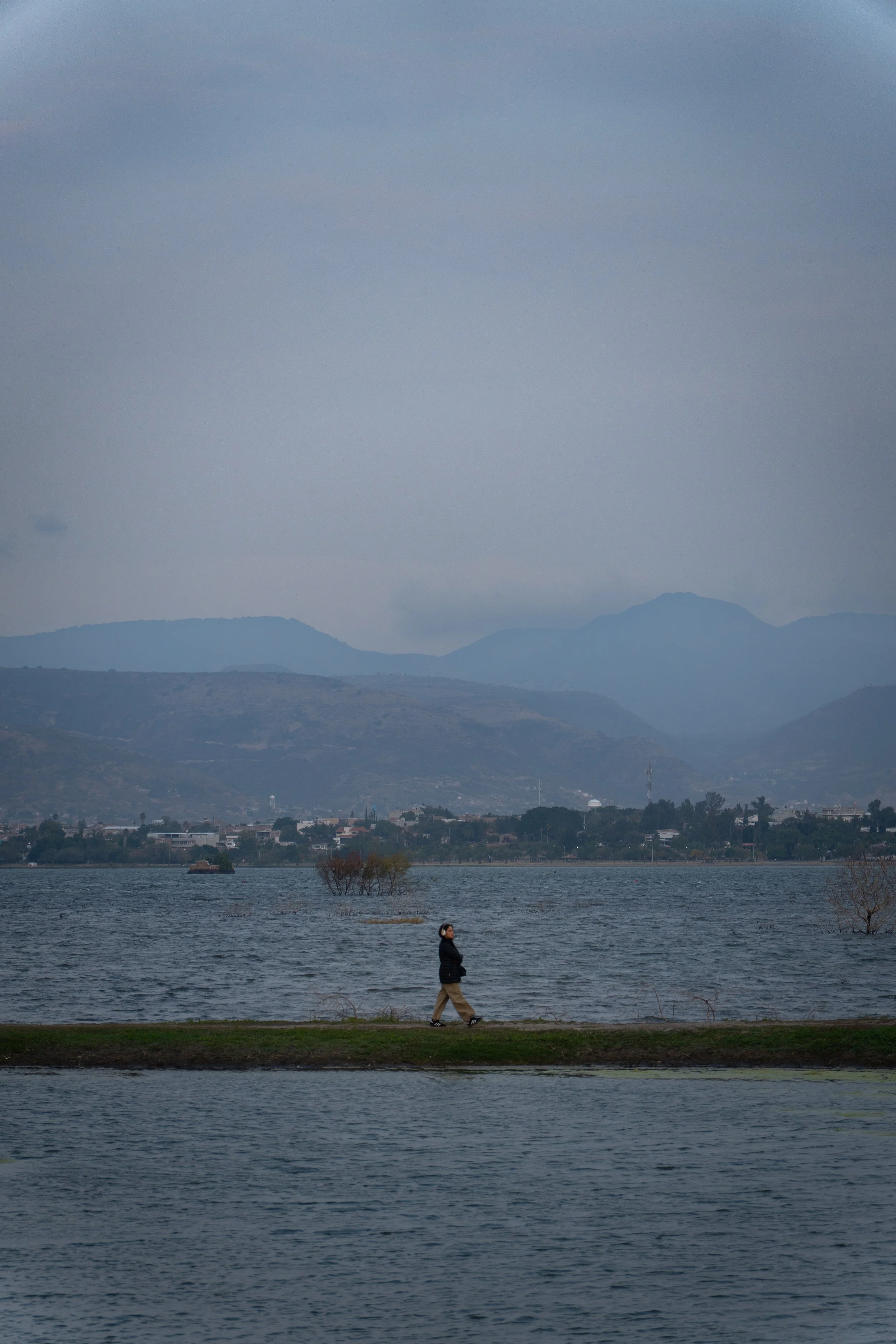 Persona caminando sola a la orilla de un lago en un día nublado. (c) 2026 Héctor Obregón