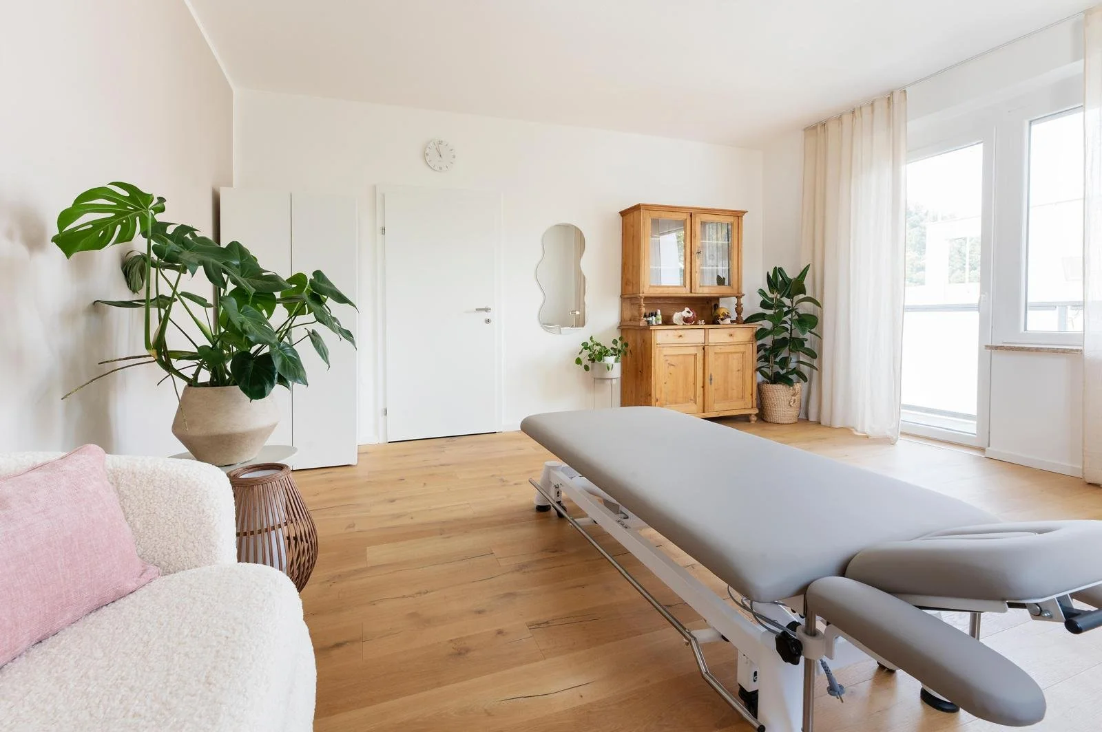 Ein heller, gemütlicher Raum mit einem Behandlungstisch, Sofas, Pflanzen und einem Holzschrank.