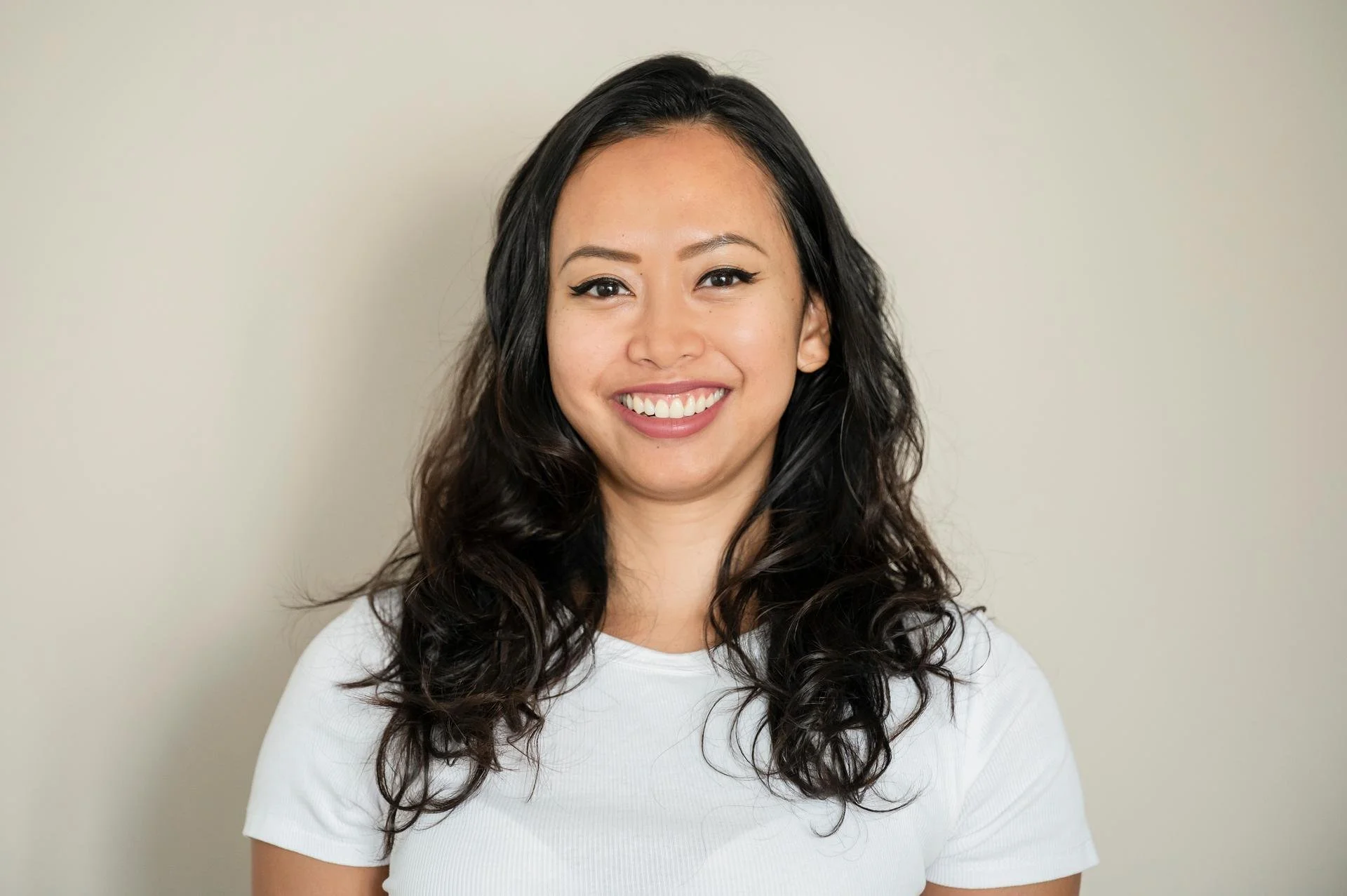 Lächelnde Frau mit schwarzen, lockigen Haaren in weißem T-Shirt vor hellgrauem Hintergrund.