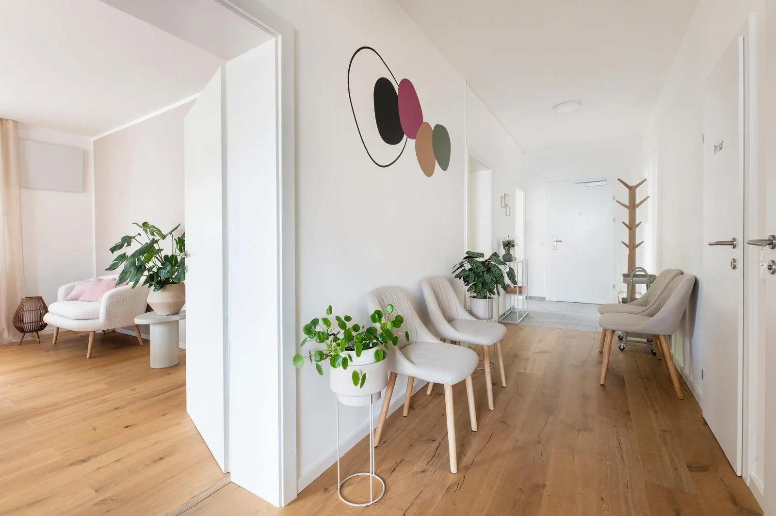 Heller, moderner Wartebereich in einer Arztpraxis mit weißen Wänden, Holzfußboden und mehreren beige Sitzstühlen. Es gibt grüne Pflanzen und eine abstrakte Wandkunst.