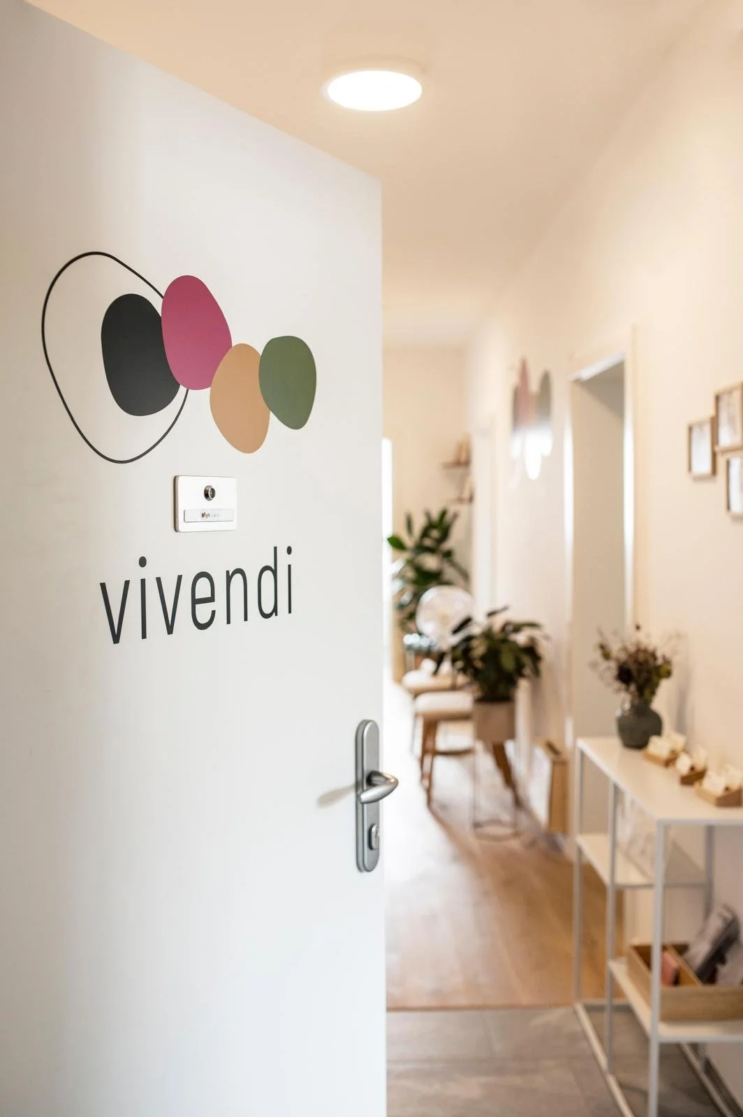 Offen gehende Tür mit dem Logo 'vivendi' und bunten ovalförmigen Designs, Blick in ein helles, dekoratives Zimmer mit Pflanzen und Bildern an der Wand.