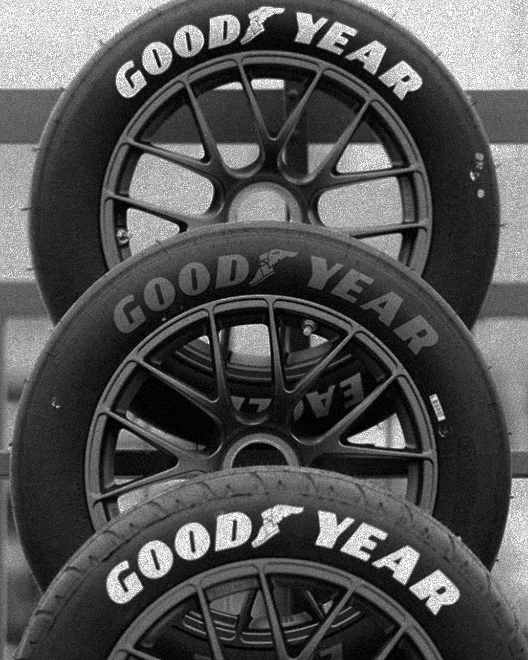 Three Goodyear tires stacked vertically with a racing wheel in the background, all on a rack.