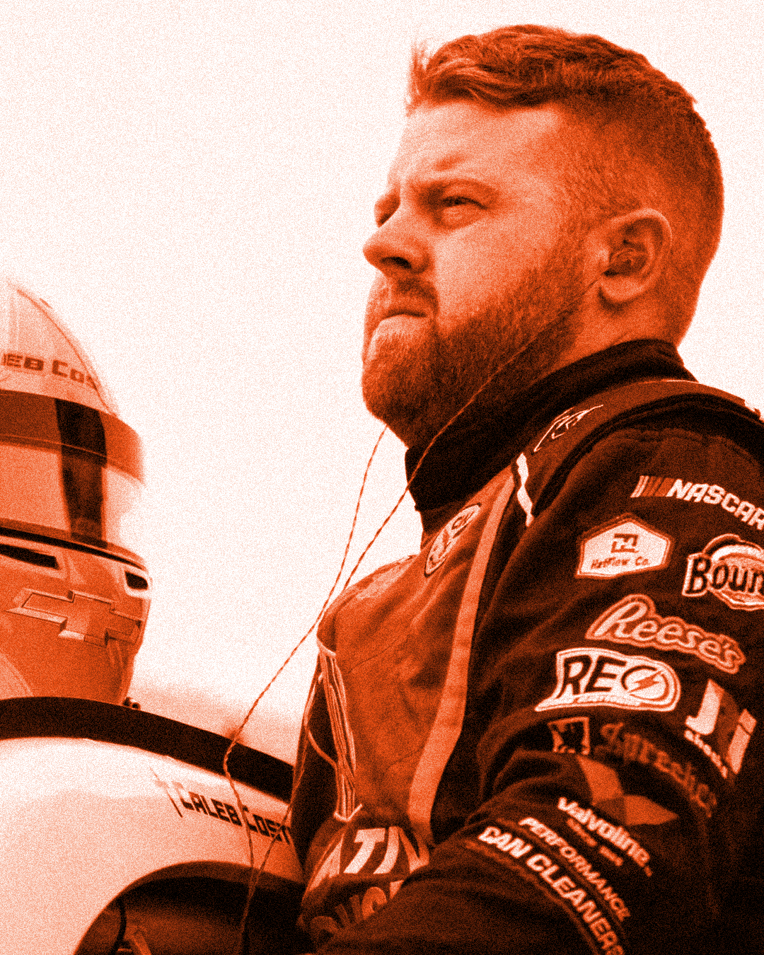 A race car driver with a beard, wearing a black racing suit with multiple sponsor logos, standing next to a helmet.