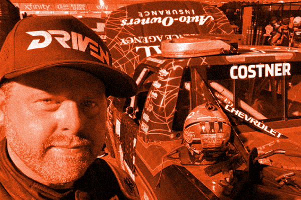 A man wearing a black cap with red and white text and a racing suit takes a selfie near a Chevrolet race car with the name 'COSTNER' on the window. The car has a helmet on the dashboard and a number 23 on the windshield.