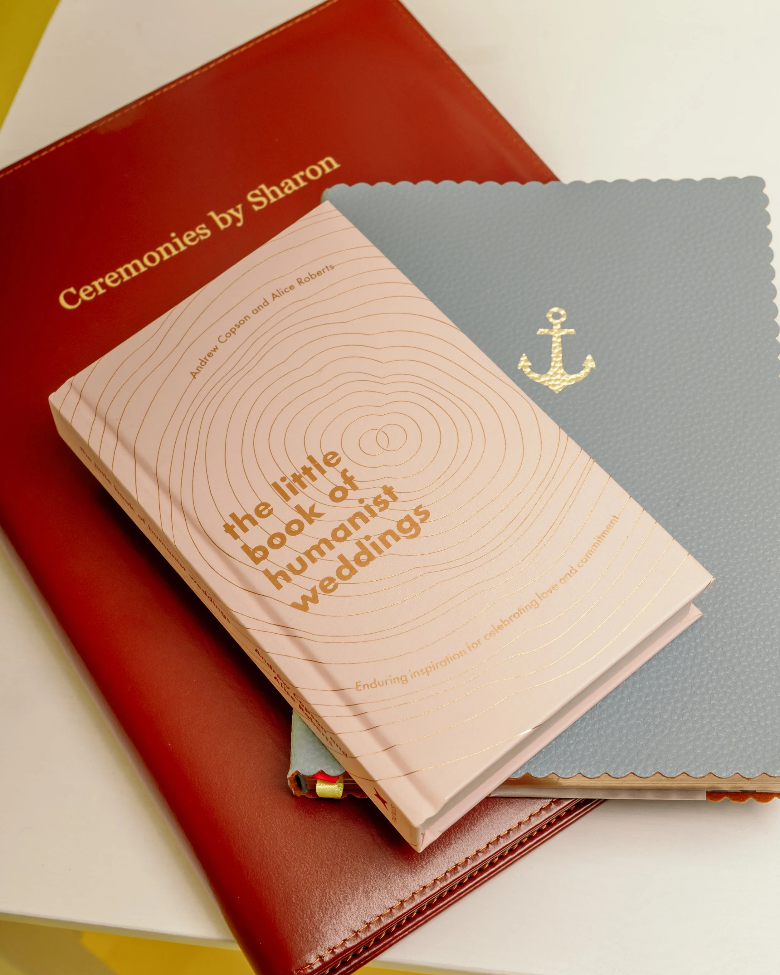 A pink book titled 'the little book of humanist weddings' resting on a blue folder with a gold anchor emblem, placed on a brown leather-bound book.