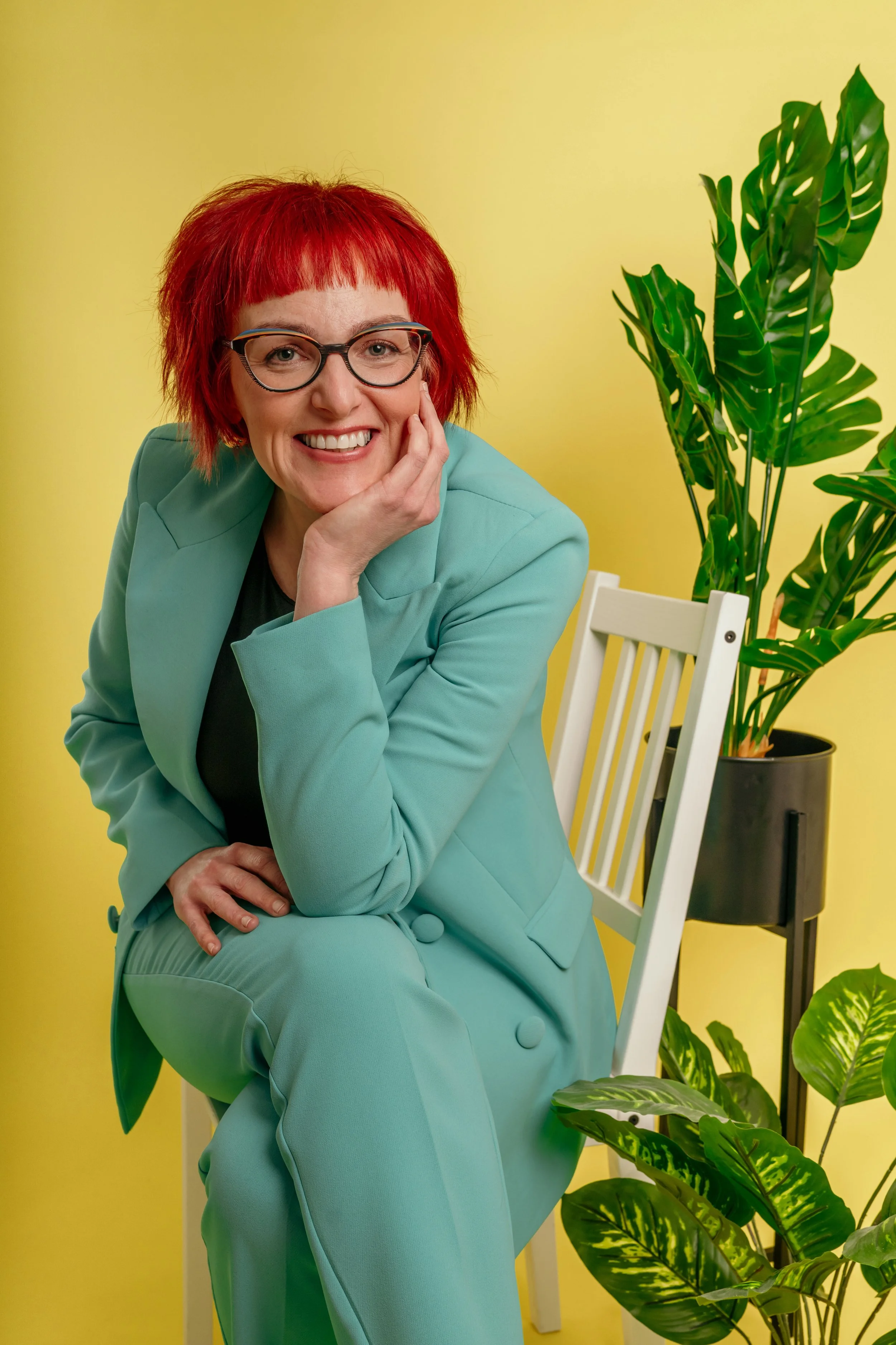 A woman with bright red hair and glasses smiling at the camera, sitting on a white chair with her hand resting on her face, wearing a mint green suit, against a yellow background with green houseplants.