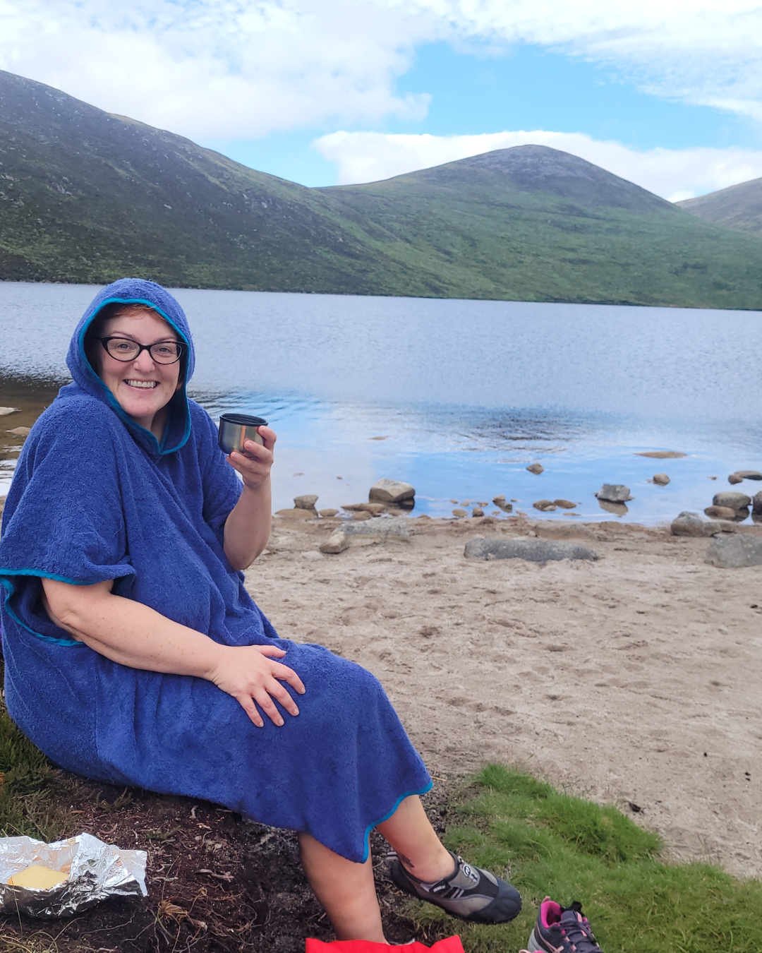 Woman sitting on grassy patch by a lake, wrapped in a blue towel with a hood, smiling, holding a metal cup, surrounded by mountains and cloudy sky.