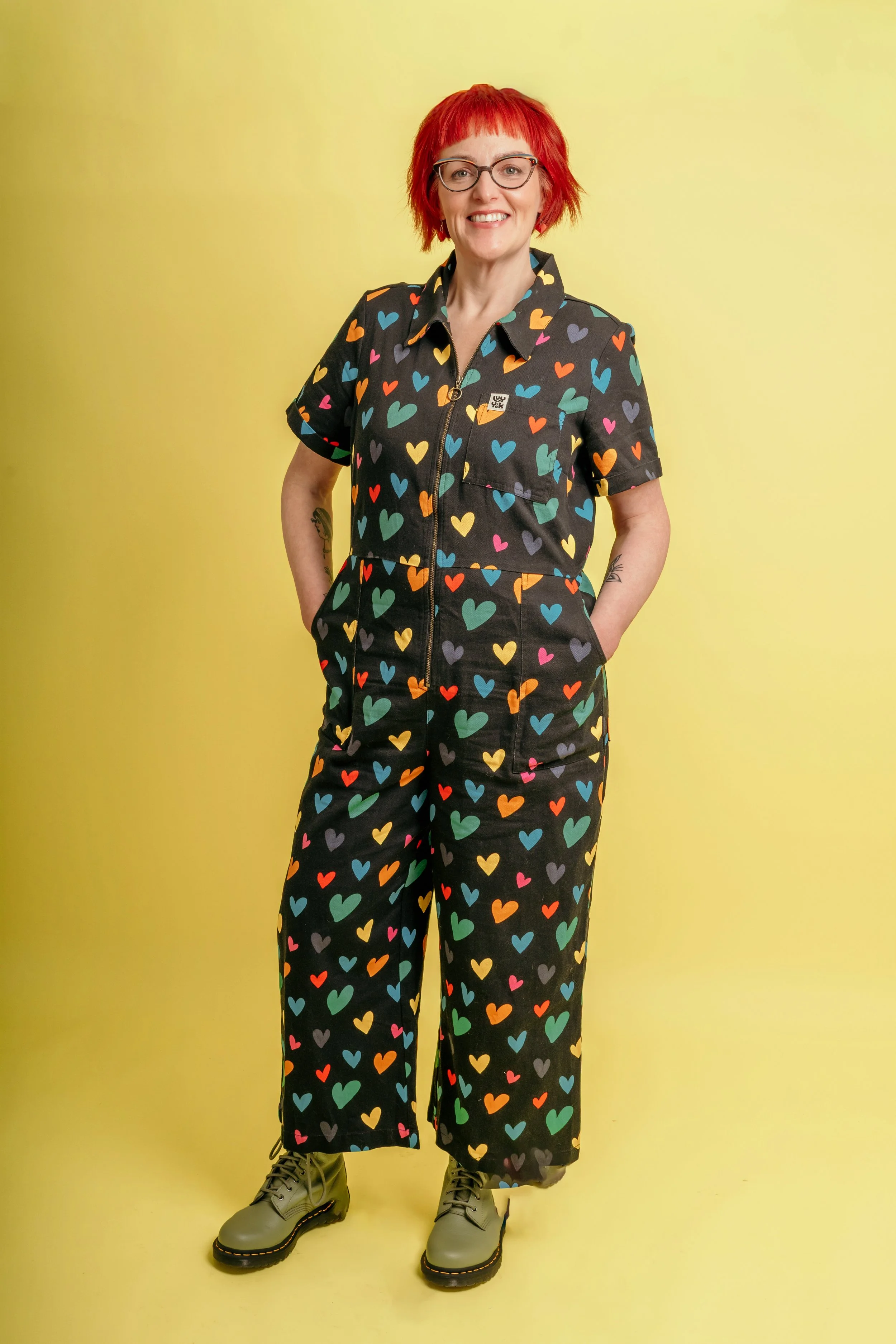 Woman with red hair and glasses wearing a black jumpsuit with colorful heart patterns, standing against a yellow background.