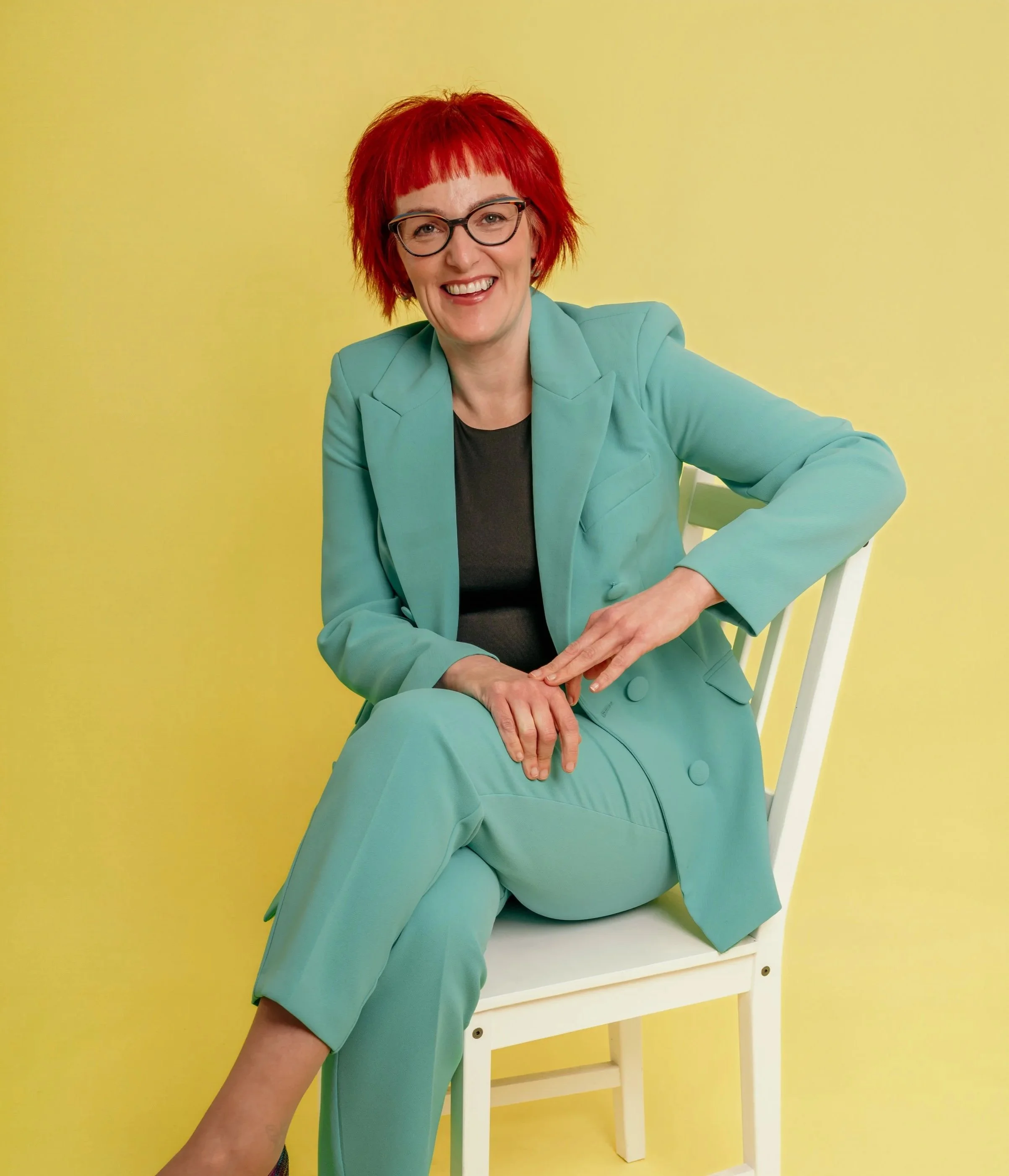 A woman with red hair and glasses sitting on a white chair against a yellow background, smiling, wearing a teal suit and black top.