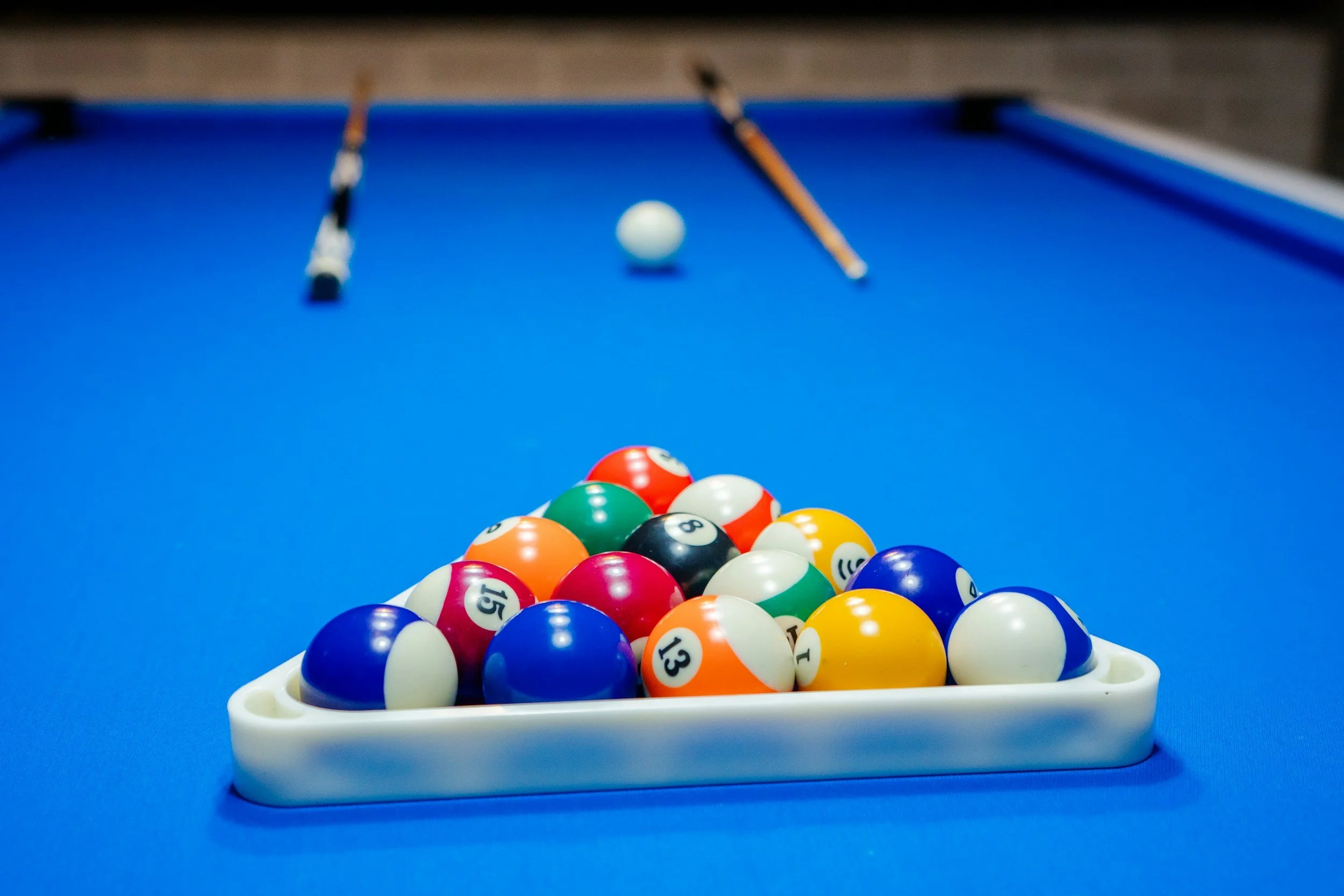 Set of pool balls racked on a blue pool table with two pool cues and the cue ball in the background.