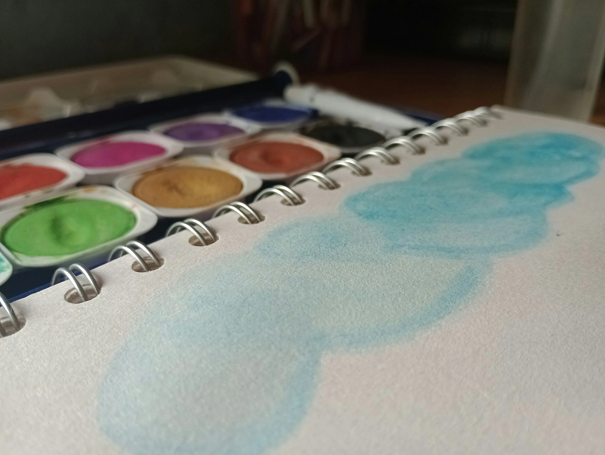 Watercolor paints and an open sketchbook with a light blue watercolor swatch.
