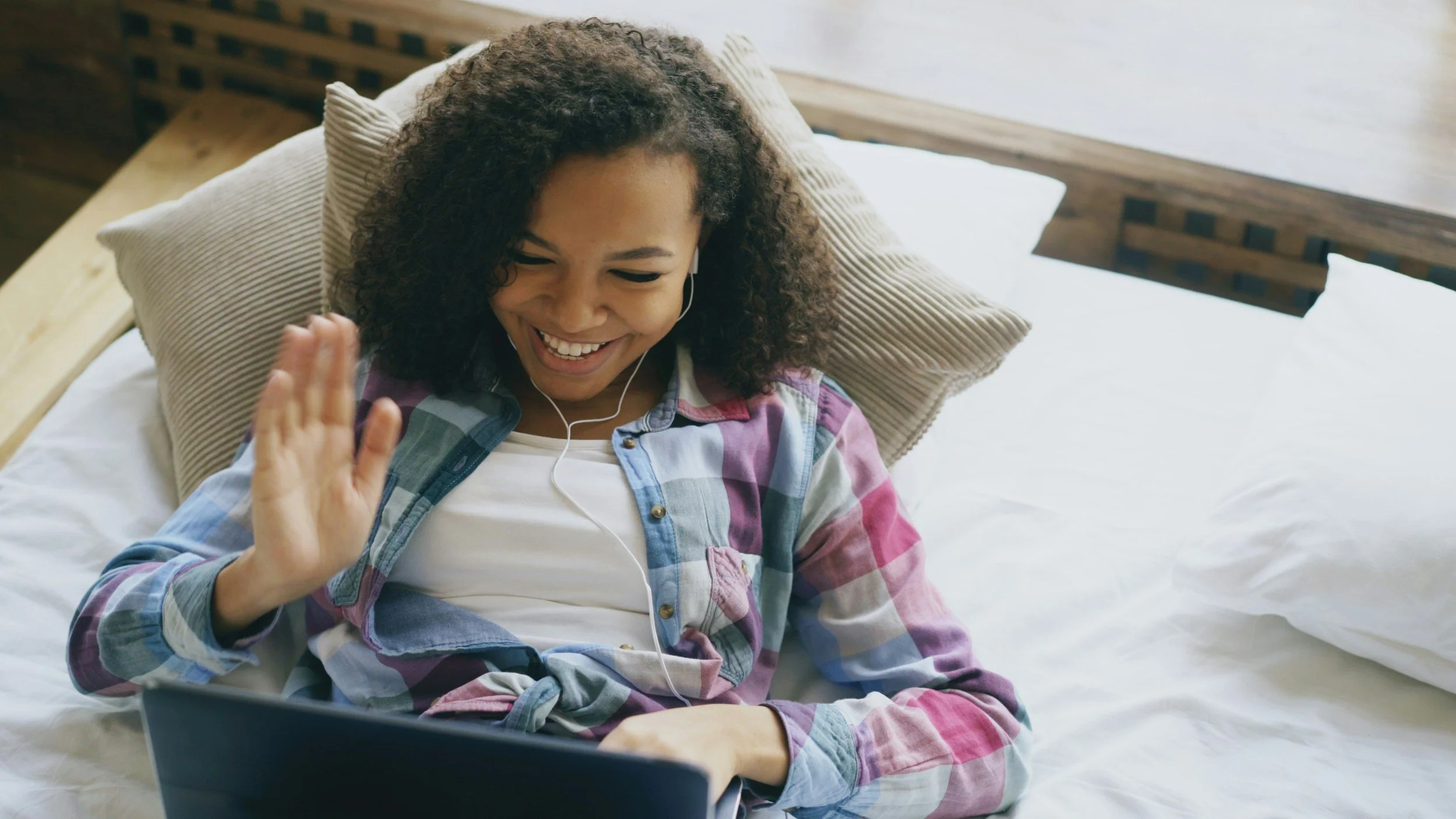 Young woman with curly hair smiling and waving while listening to headphones and watching a laptop from her bed.