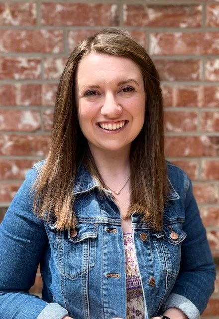 A smiling woman with long brown hair wearing a denim jacket standing in front of a brick wall.