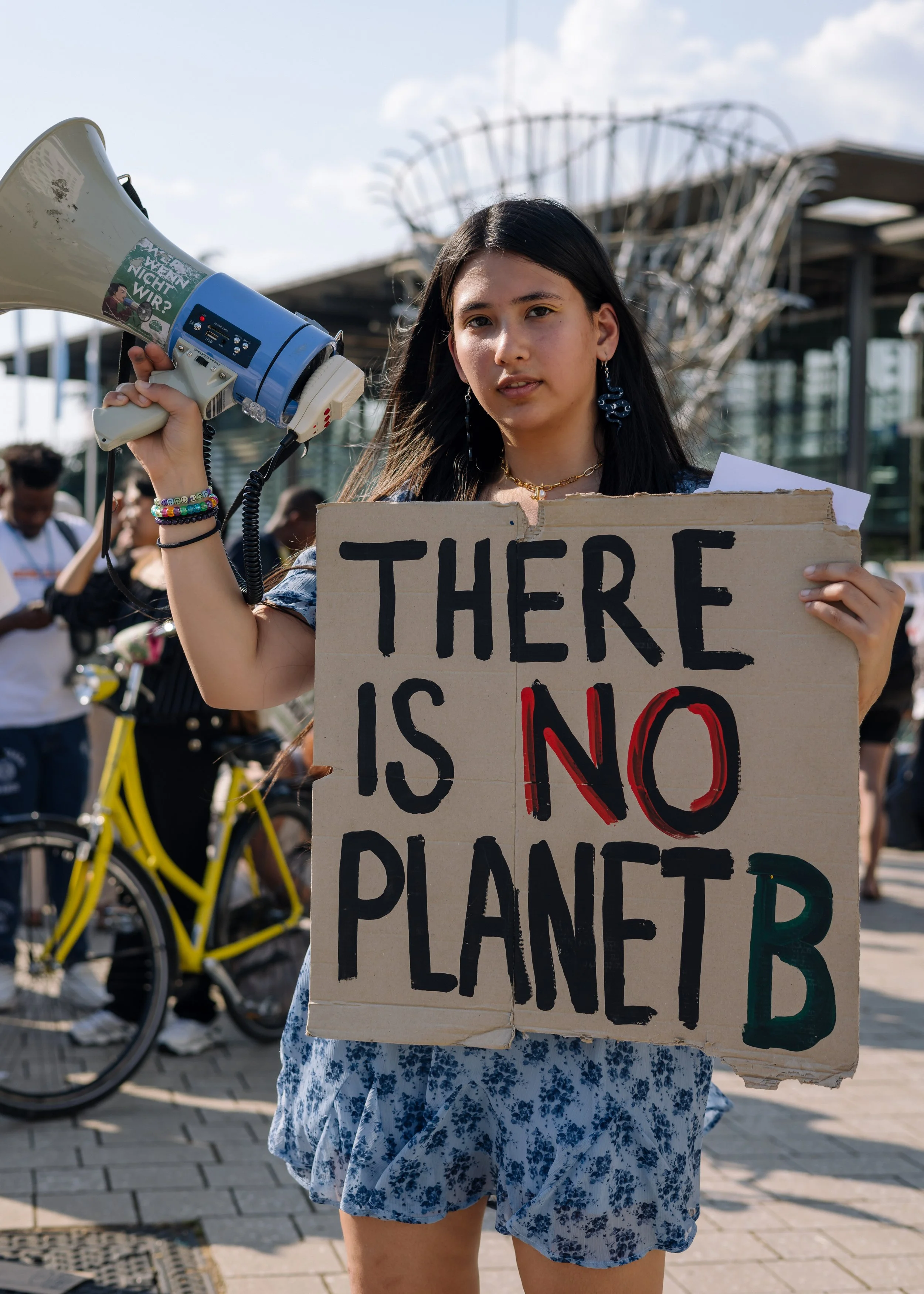 Alexia Leclercq young woman holding a cardboard sign that reads 'There is no planet B' and a megaphone during a protest or demonstration, with other people and bicycles in the background.