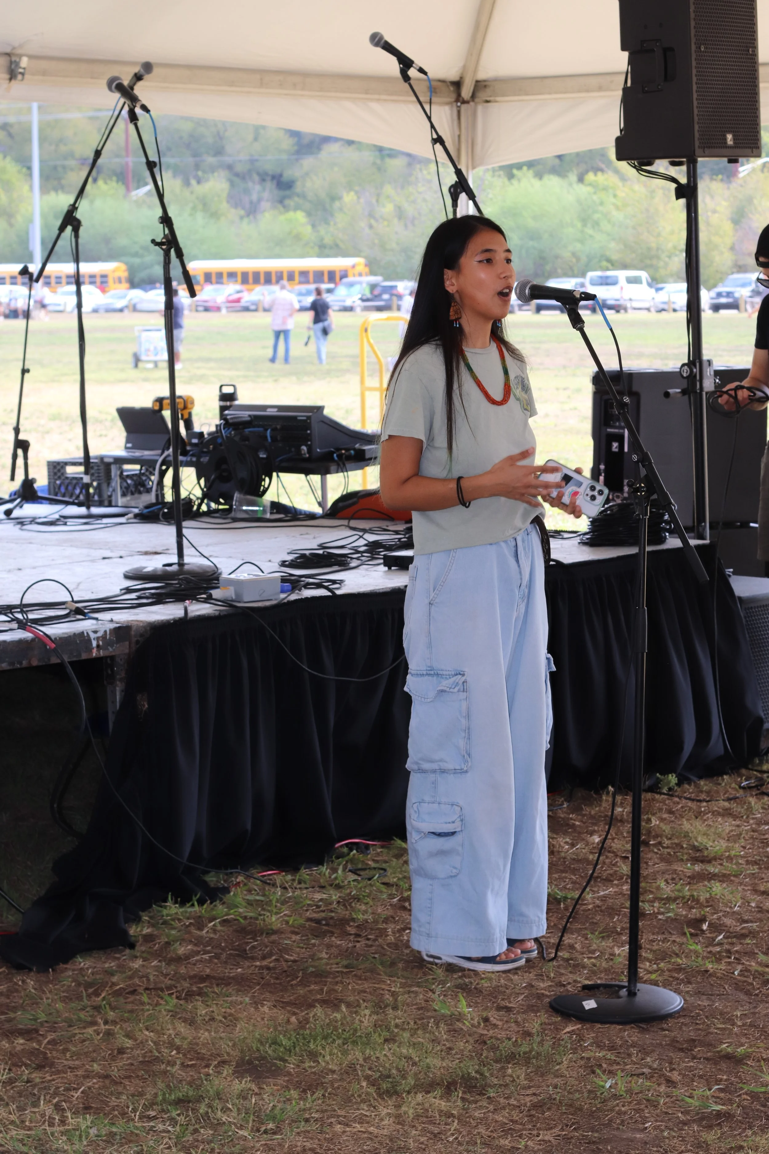 A young woman is speaking into a microphone on a stage at an outdoor event. She is holding a device in her hands. The stage has sound equipment, microphones, and cables. There is a tent overhead, and in the background, there are parked cars, a school bus, and trees.