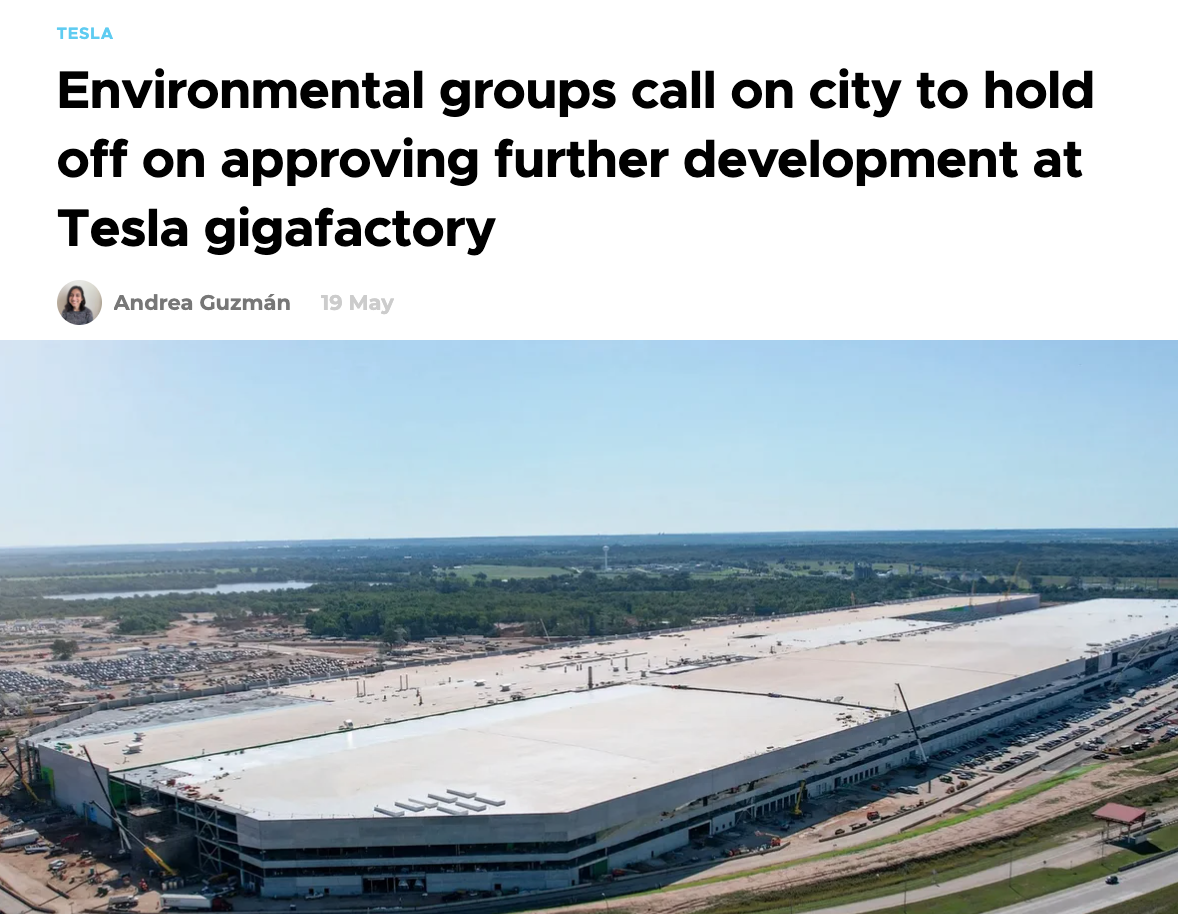Aerial view of Tesla gigafactory with a white roof and parking lot surrounding it, set in a green landscape under a clear blue sky.