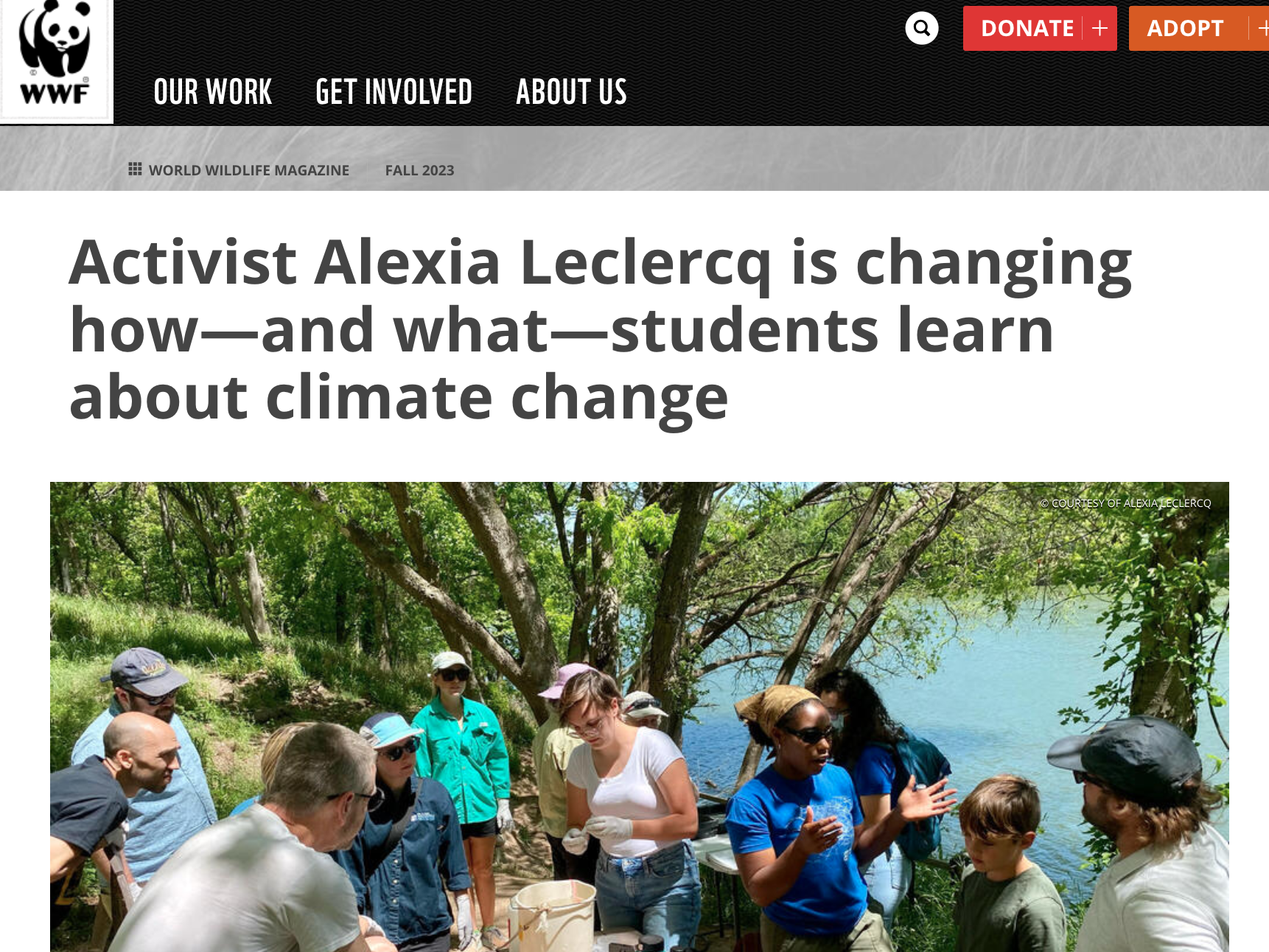 A group of people, including children and adults, gathered outdoors near a body of water surrounded by green trees, listening to a woman in a blue shirt speak during a natural education or environmental activity.