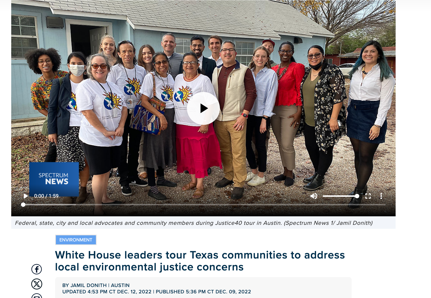 Group of diverse people, including men and women, standing outdoors in front of a blue house, smiling for a photo. Some are wearing white t-shirts with a logo, and others are dressed in casual and business casual attire.