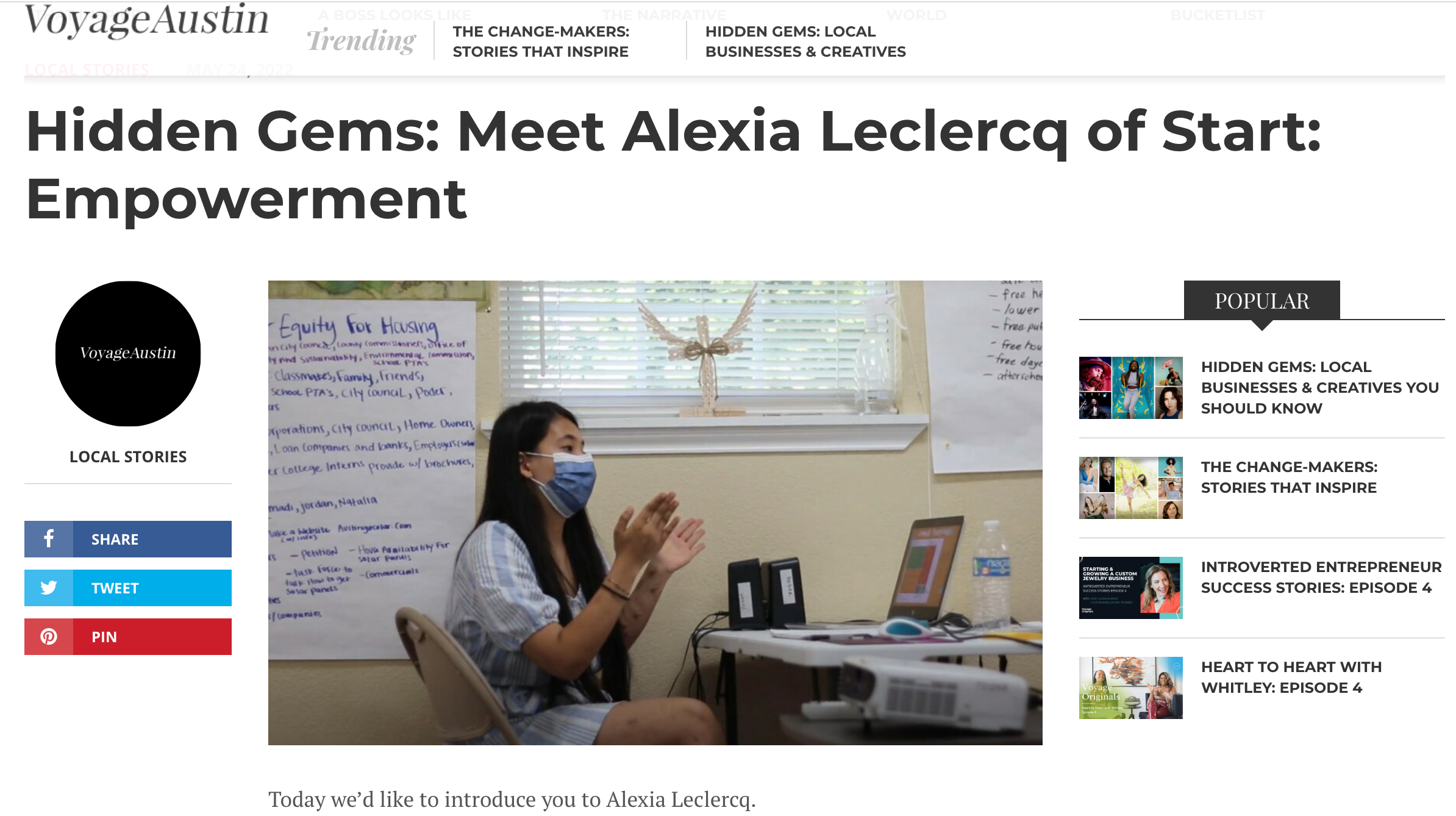 A woman with long dark hair, wearing a blue face mask and a striped shirt, sits at a desk with a laptop and a water bottle in a room with beige walls and a large window with blinds. Behind her, there is a poster on the wall with handwritten notes tit