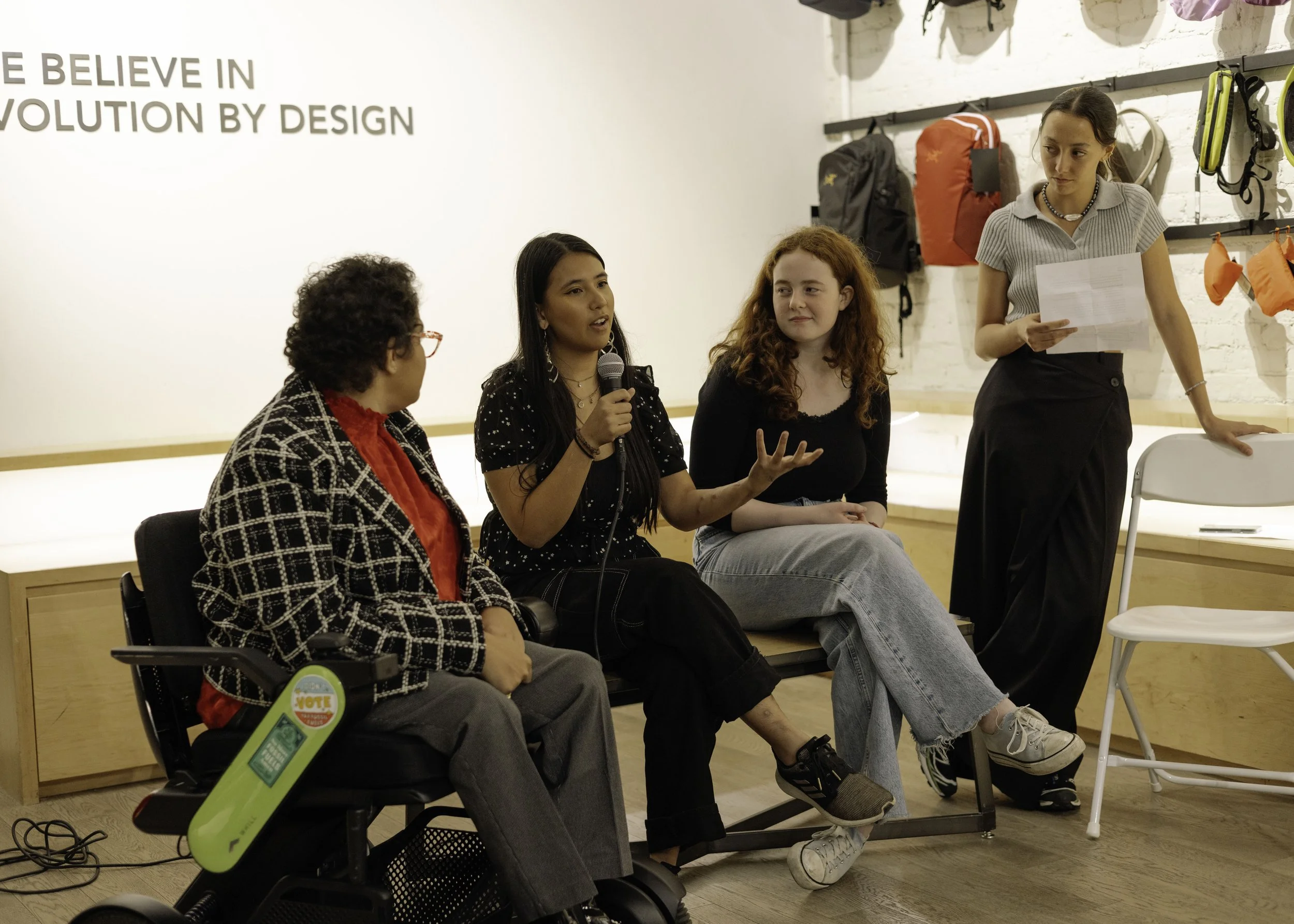 Four women engaging in a discussion in a retail or community space with backpacks and gear on the wall behind them.