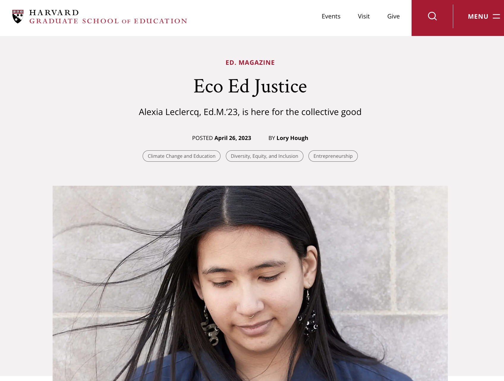 A young woman with long black hair, earrings, and a septum piercing, looking down with her eyes closed, standing in front of a light gray, textured stone wall.