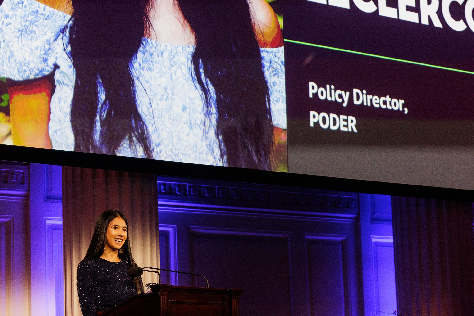 Young woman with long dark hair speaking at a podium during a presentation, with a large screen behind her displaying her name, 'Policy Director, PODER', and a partial picture of her.