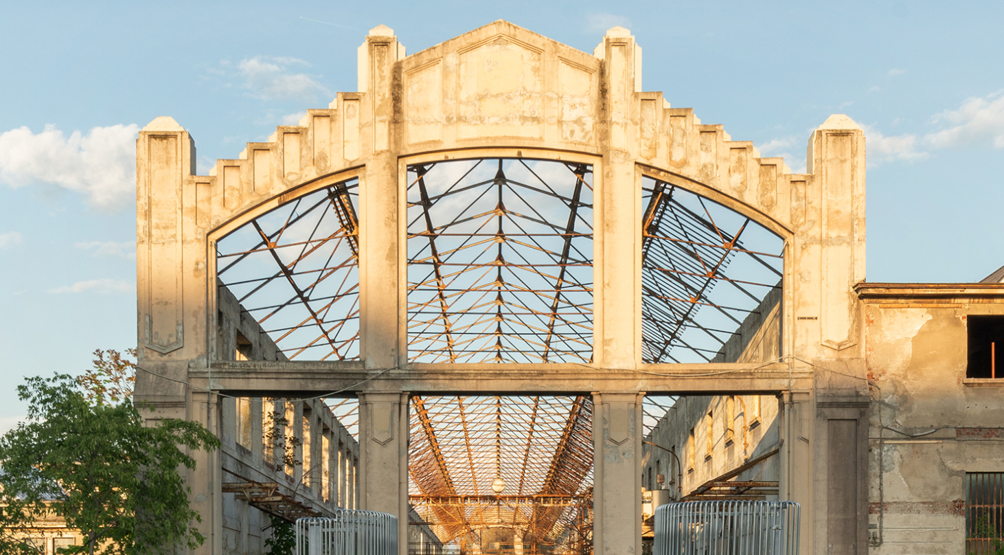 An abandoned industrial building with an arch-shaped entrance and a rusted iron framework inside, during daylight.