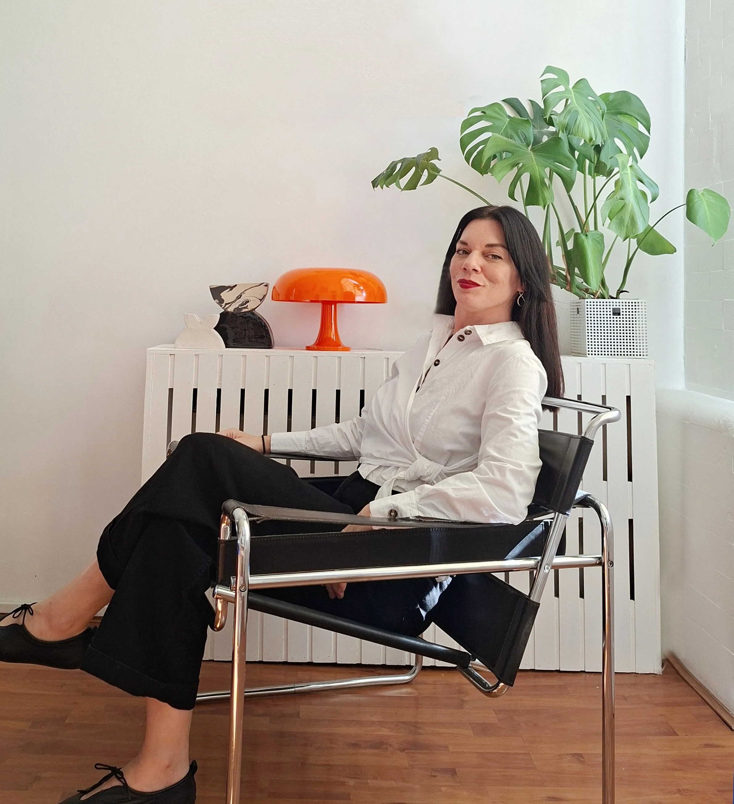 Woman sitting in a chair with one leg crossed over the other, wearing a white shirt and black pants, with a large green plant behind her and decorative objects on a white table to her side.