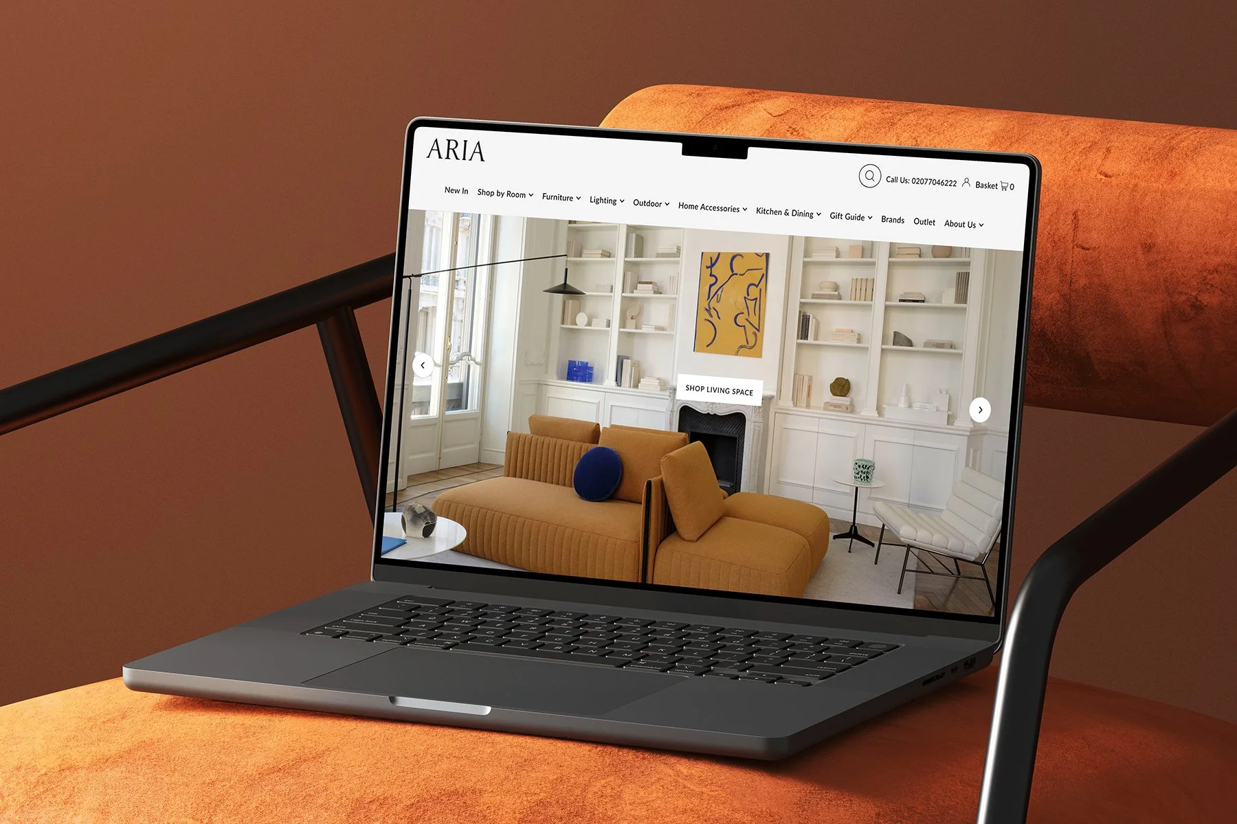 A laptop showing an interior decorating website with a living room featuring white shelves, a fireplace, tan sofa, white chair, and artwork, resting on an orange surface with a black chair in the foreground.