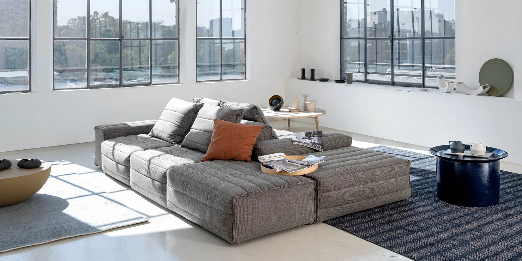 Modern living room with a large gray sectional sofa, decorative pillows, a black round coffee table with carafes, and a light wood side table with ceramics, against large windows revealing an urban cityscape.