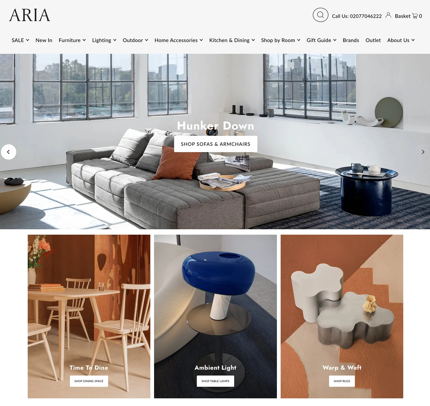 Screenshot of ARIA home furniture website homepage featuring a large modern living room with gray sectional sofa, orange pillow, and coffee table. Below are images of dining chairs, a blue table lamp, and abstract rugs, with shopping prompts.
