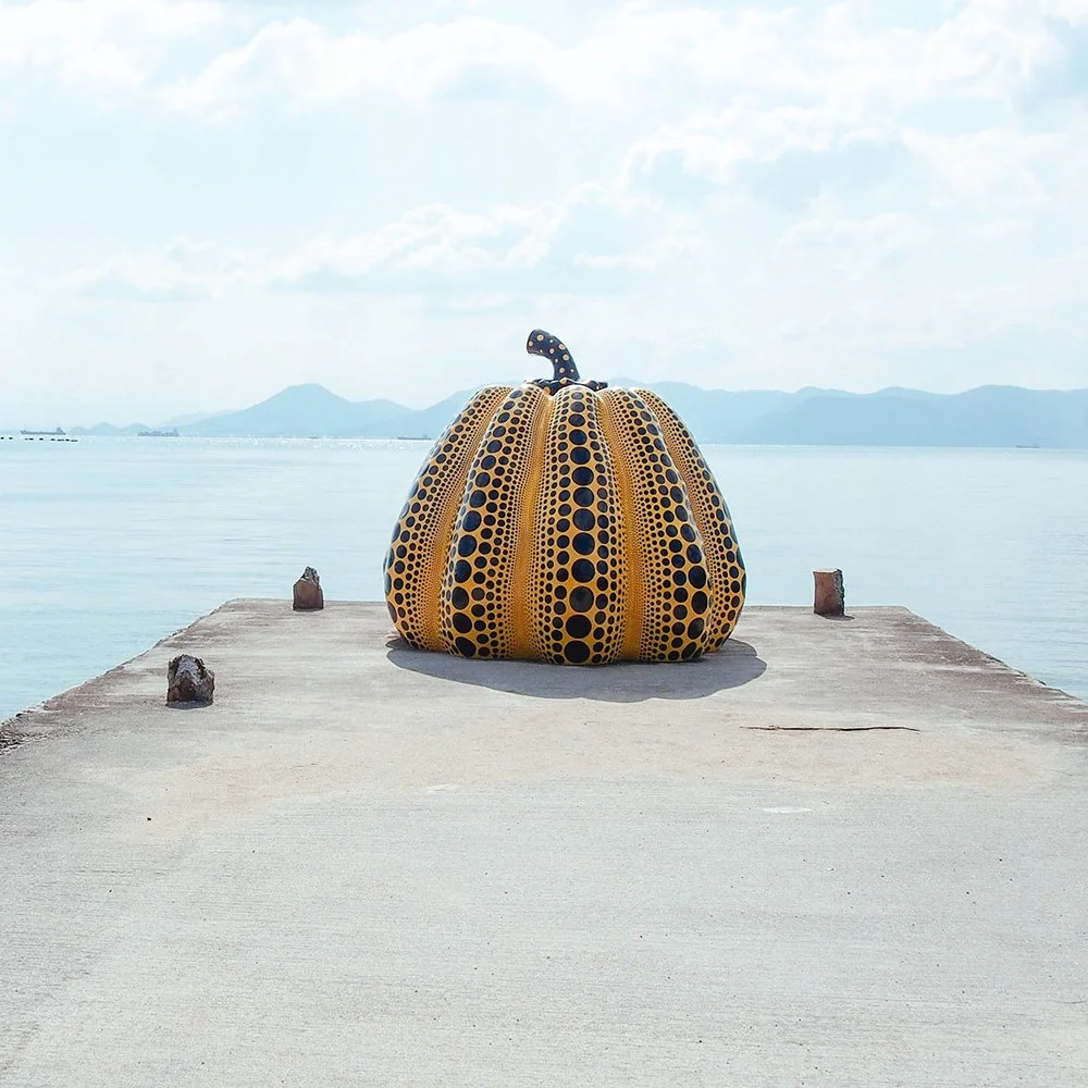 Large pumpkin sculpture with yellow and black dotted pattern on a concrete pier by a body of water, with mountains and cloudy sky in the background.