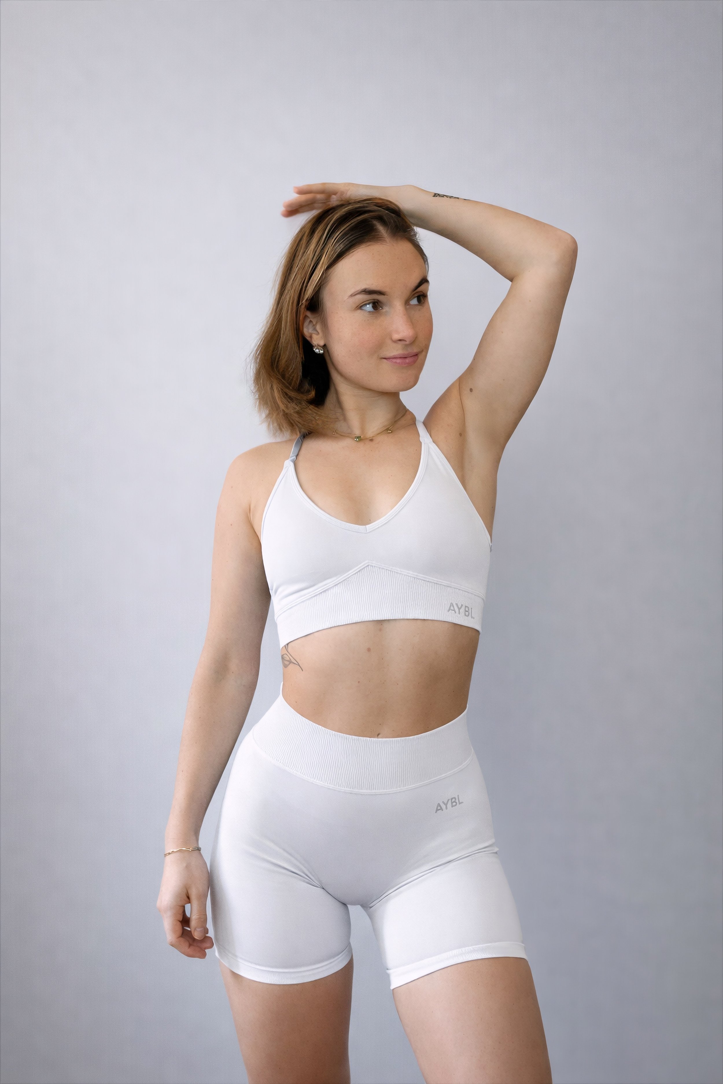 Junge Frau in weißem Sportoutfit steht vor neutralem Hintergrund, hebt ihren Arm und schaut nach rechts.