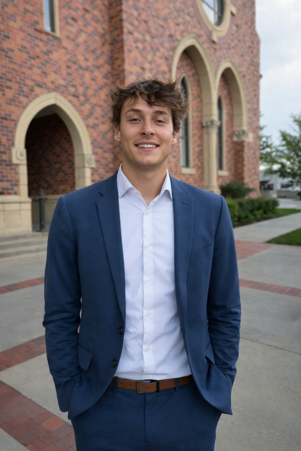A young man in a blue suit and white shirt standing outside in front of a brick building with arched windows.
