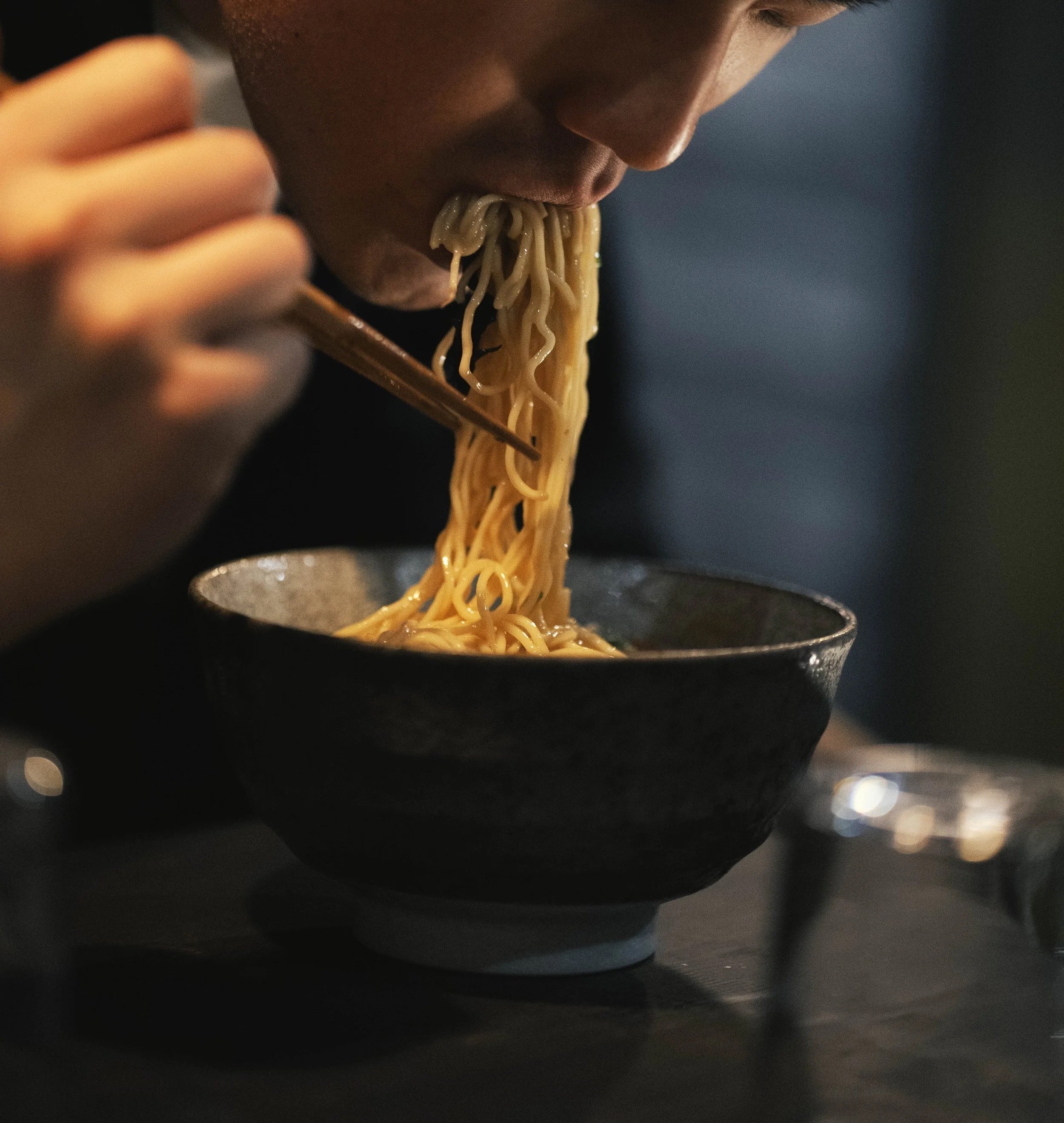Persona che mangia ramen con dei chopstick in un ambiente buio.