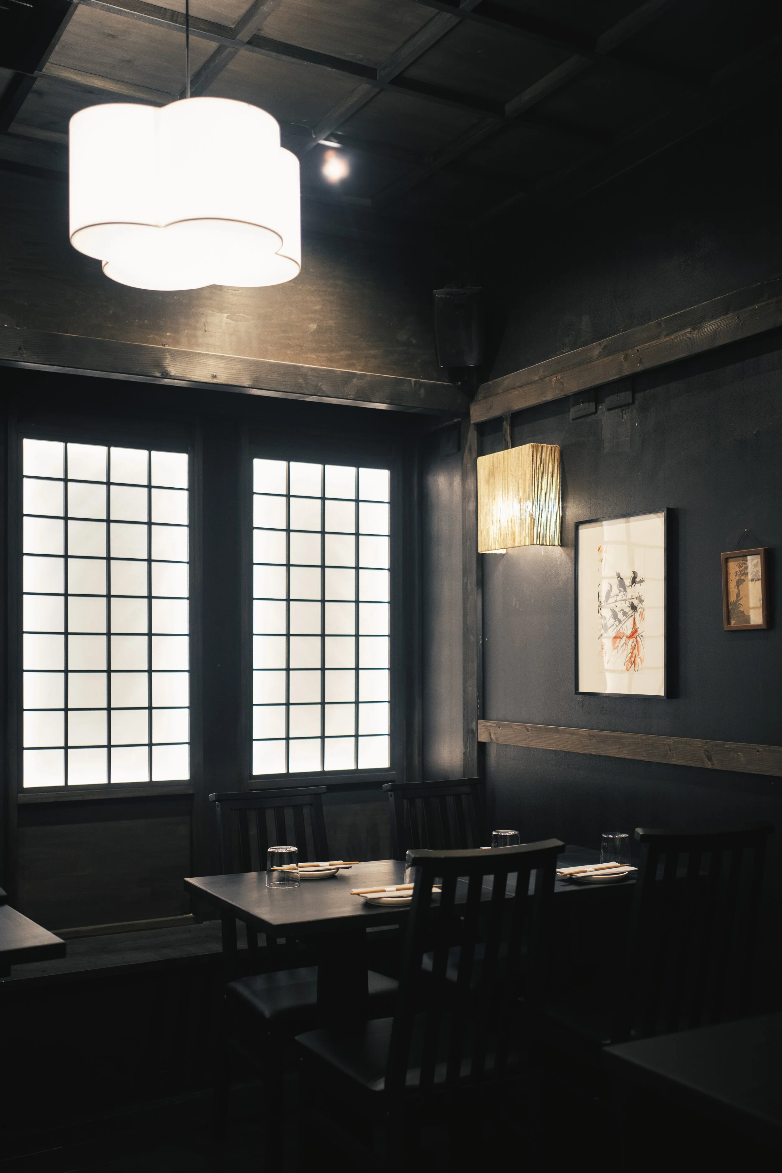 Interno di un ristorante con pareti scure e decorazioni minimaliste, tavolo pronto con piatti e bicchieri, finestre con infissi in stile shoji, illuminazione calda. 