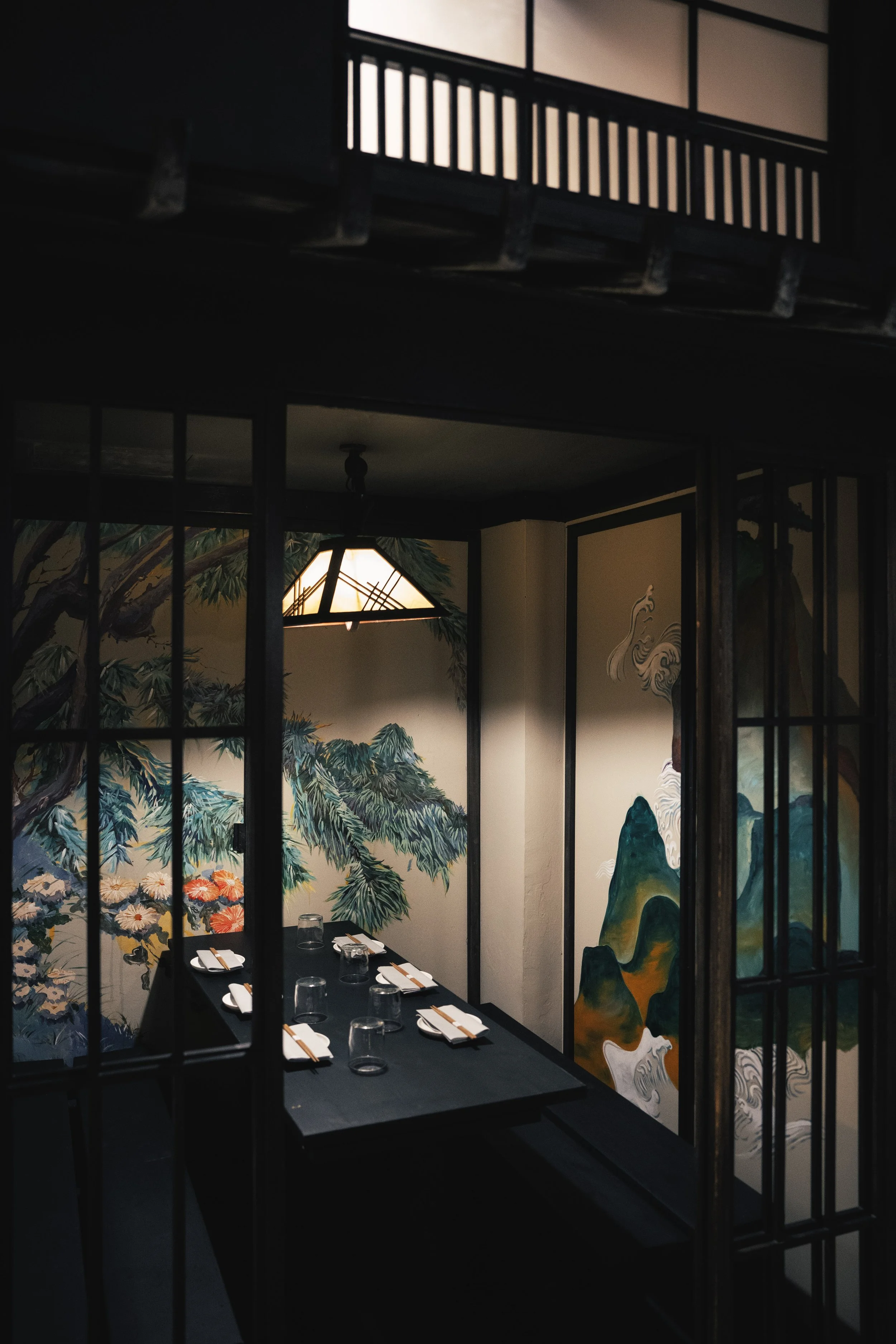 Sala da pranzo con pareti decorate con dipinti di paesaggi geografici e naturali, vista attraverso una porta in stile giapponese, tavolo apparecchiato con piatti, bicchieri e bacchette.