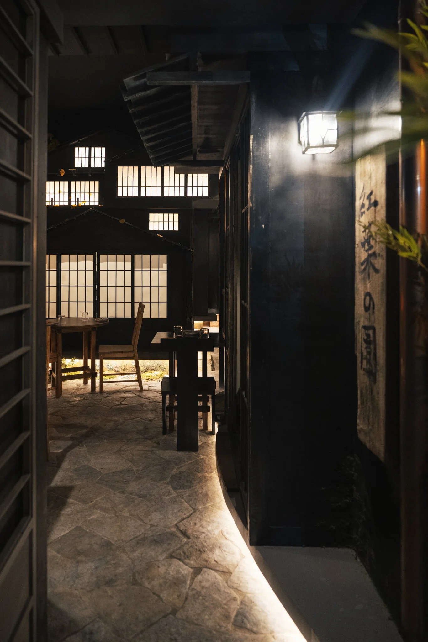 Interno di un ristorante con pareti nere, tavoli di legno e luce soffusa, con finestre in stile giapponese.