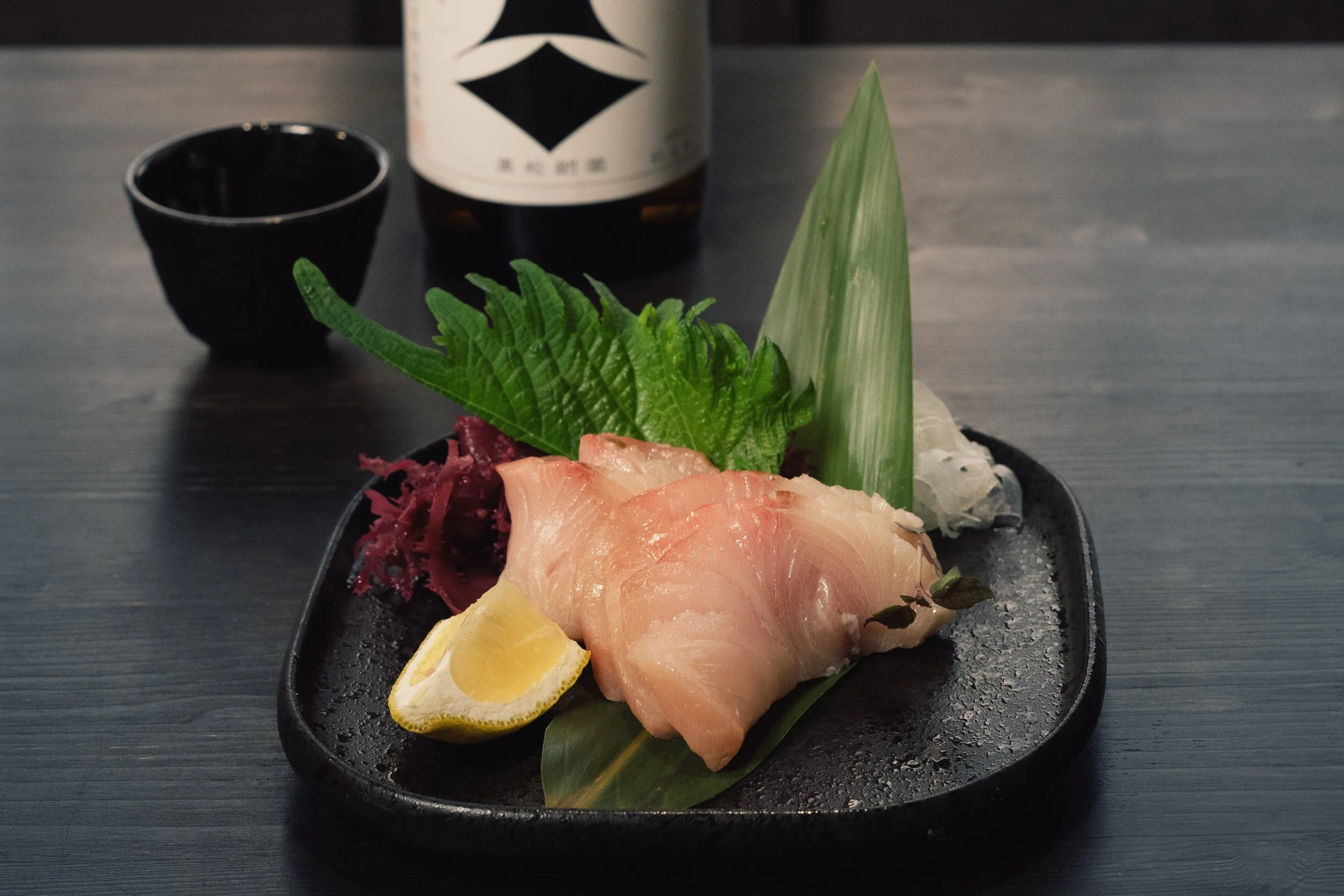 Fetta di sashimi di pesce crudo, limone, foglie di shiso e condimenti su un piatto nero, con bottiglia di sake e tazza in sfondo.