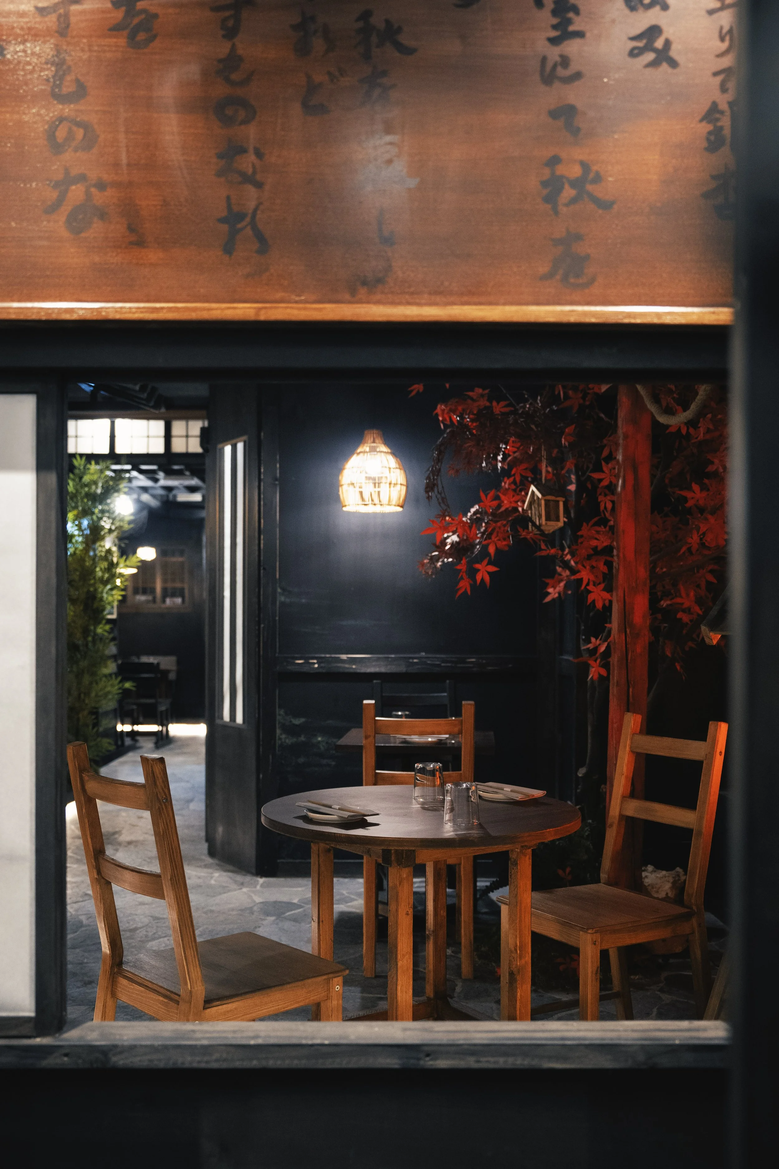 Interno di un ristorante giapponese con un tavolo rotondo in legno, tre sedie in legno, un'illuminazione calda e decorazioni che ricordano l'autunno, come foglie rosse e una pianta di acero rosso.