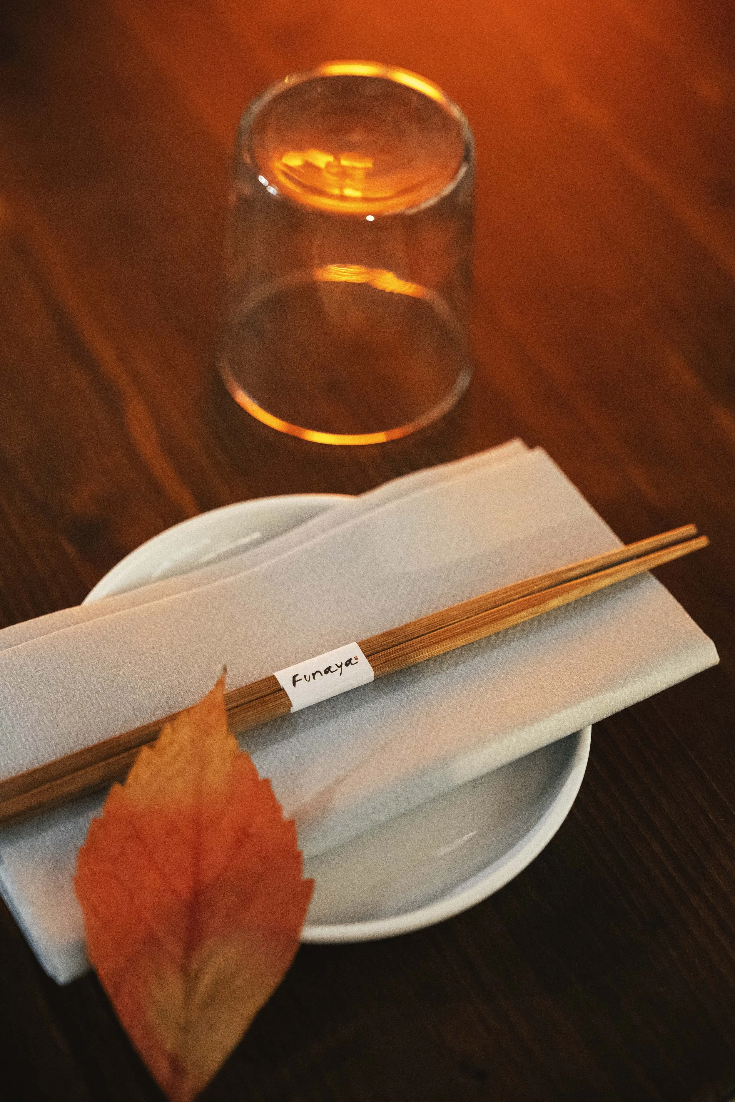 Tavolo apparecchiato con una tazza bianca, un tovagliolo di stoffa, delle bacche di bambù con un'etichetta con scritto "Funaya", un bicchiere d'acqua rovesciato e una foglia di colore arancione.