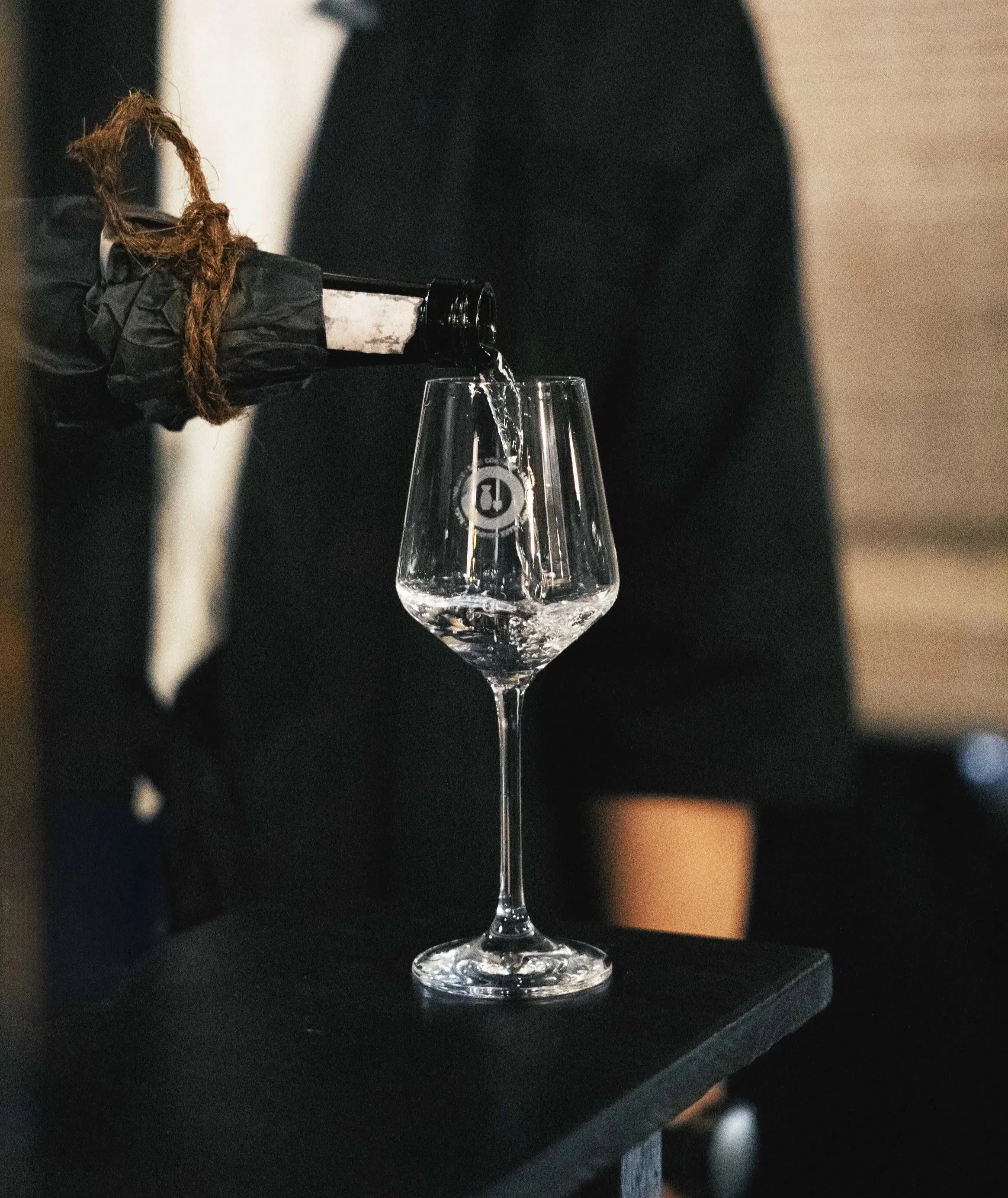 Una mano con guanti di plastica versa vino o champagne in un bicchiere di vetro su un tavolo nero.