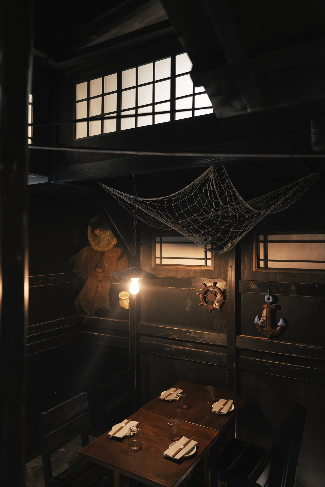 Interno di un ristorante con pareti in legno, tavolo preparato per quattro persone, illuminazione calda, decorazioni marittime come un'ancora e un timone, rete da pesca appesa al soffitto e finestra con vetri opachi.