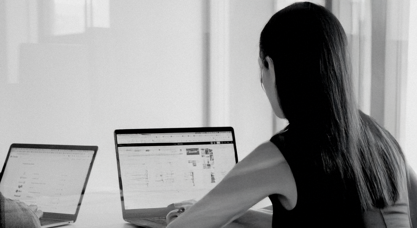 A woman with long hair sitting at a desk using two laptops, one in front of her and another to her left.