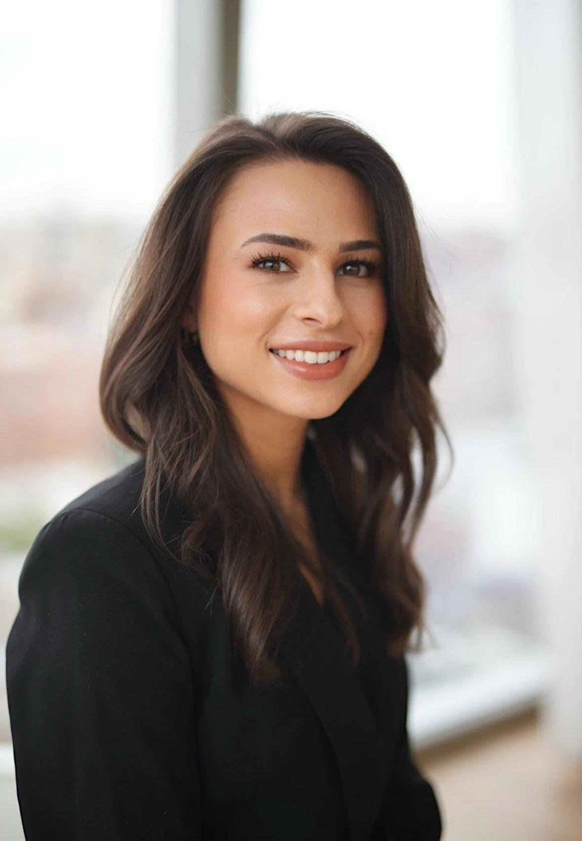 A young woman with long, wavy dark brown hair, wearing a black blazer, smiling in front of a bright window.