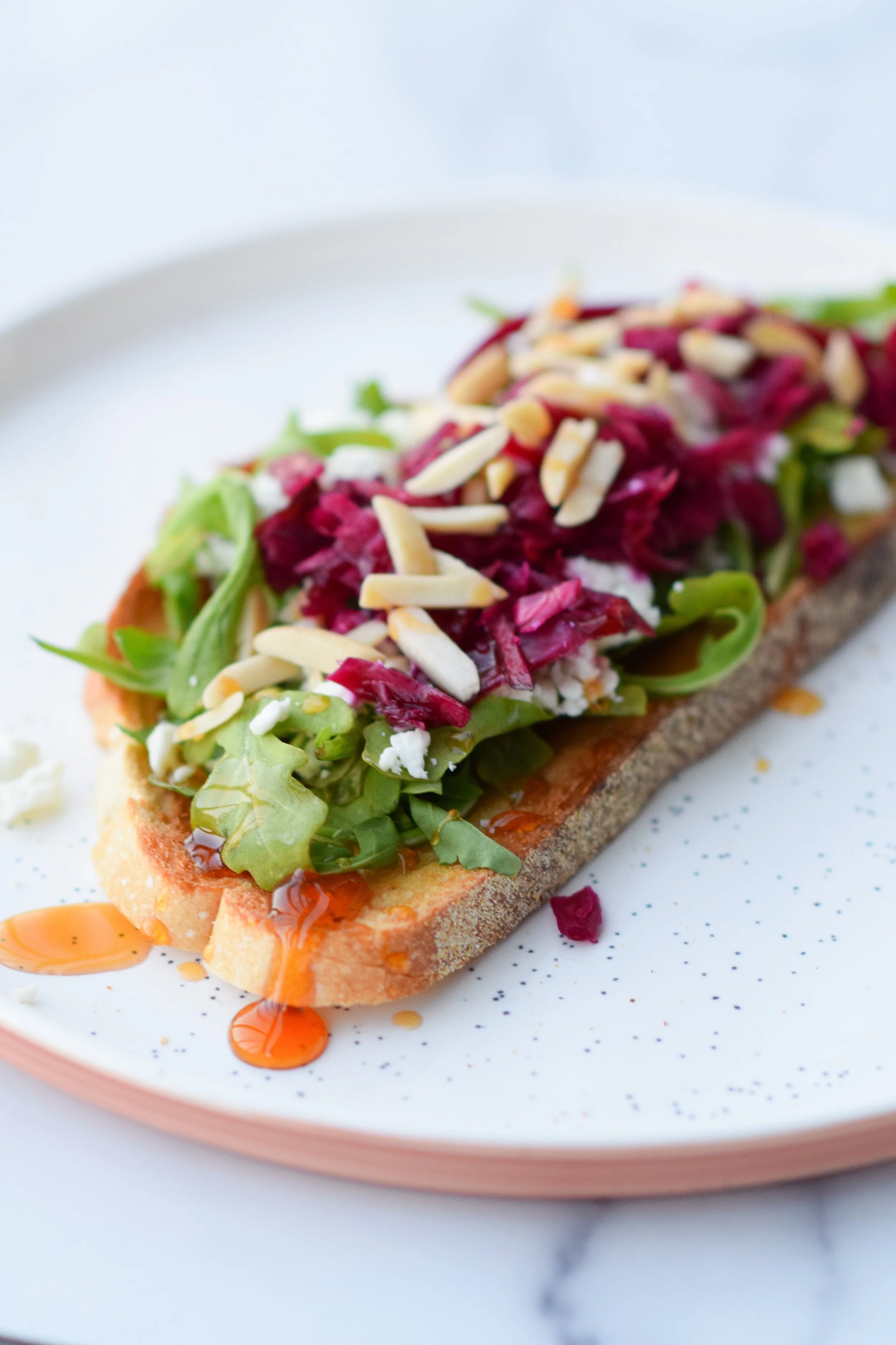 An open-faced sandwich with lettuce, shredded beets, chopped nuts, and a drizzle of orange sauce on toasted bread, served on a white speckled plate.