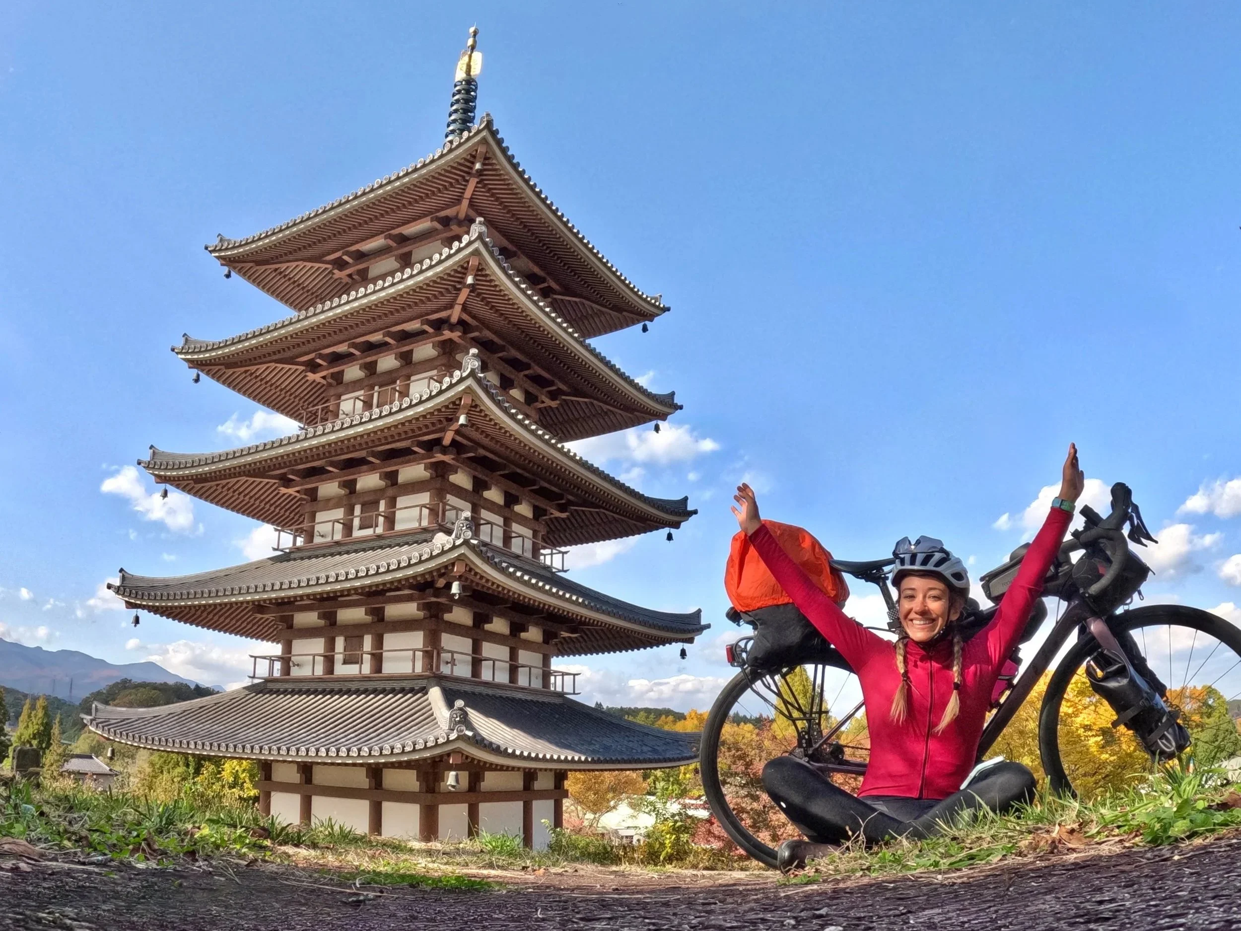 Smiling woman in a pink jacket and helmet sitting cross-legged on the ground with a mountain bike, raising her arms, in front of a traditional Japanese pagoda under a blue sky.