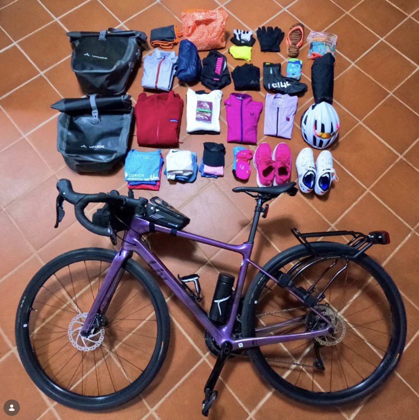 A purple Trek bicycle with a water bottle on the frame is positioned in front of a collection of various folded and packed clothes, shoes, helmet, gloves, and other cycling gear on a tiled floor.